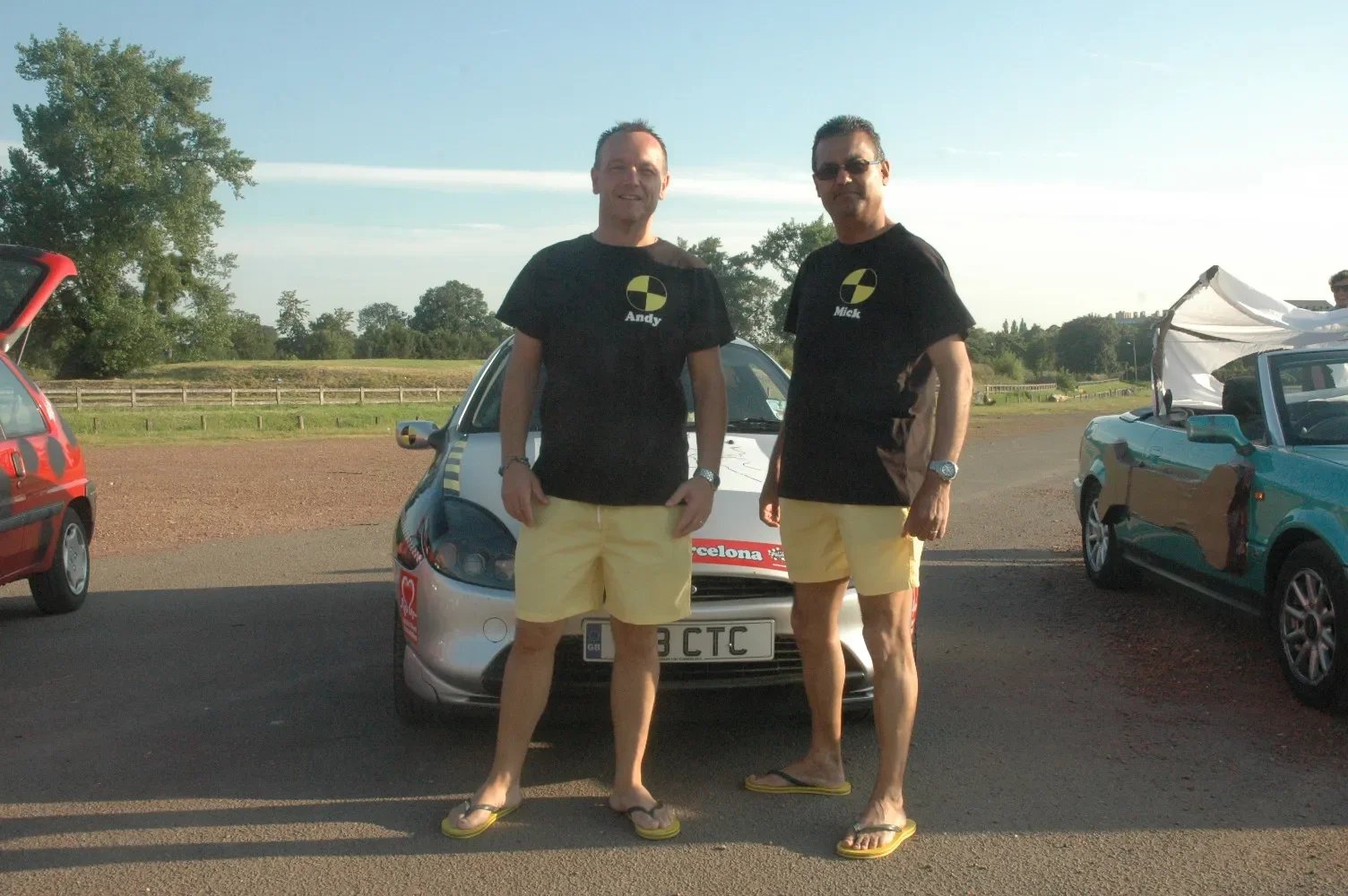 Two men wearing black T-shirts with a yellow and black logo, yellow shorts, and flip-flops standing in front of a silver car, with several other cars parked nearby and trees in the background.