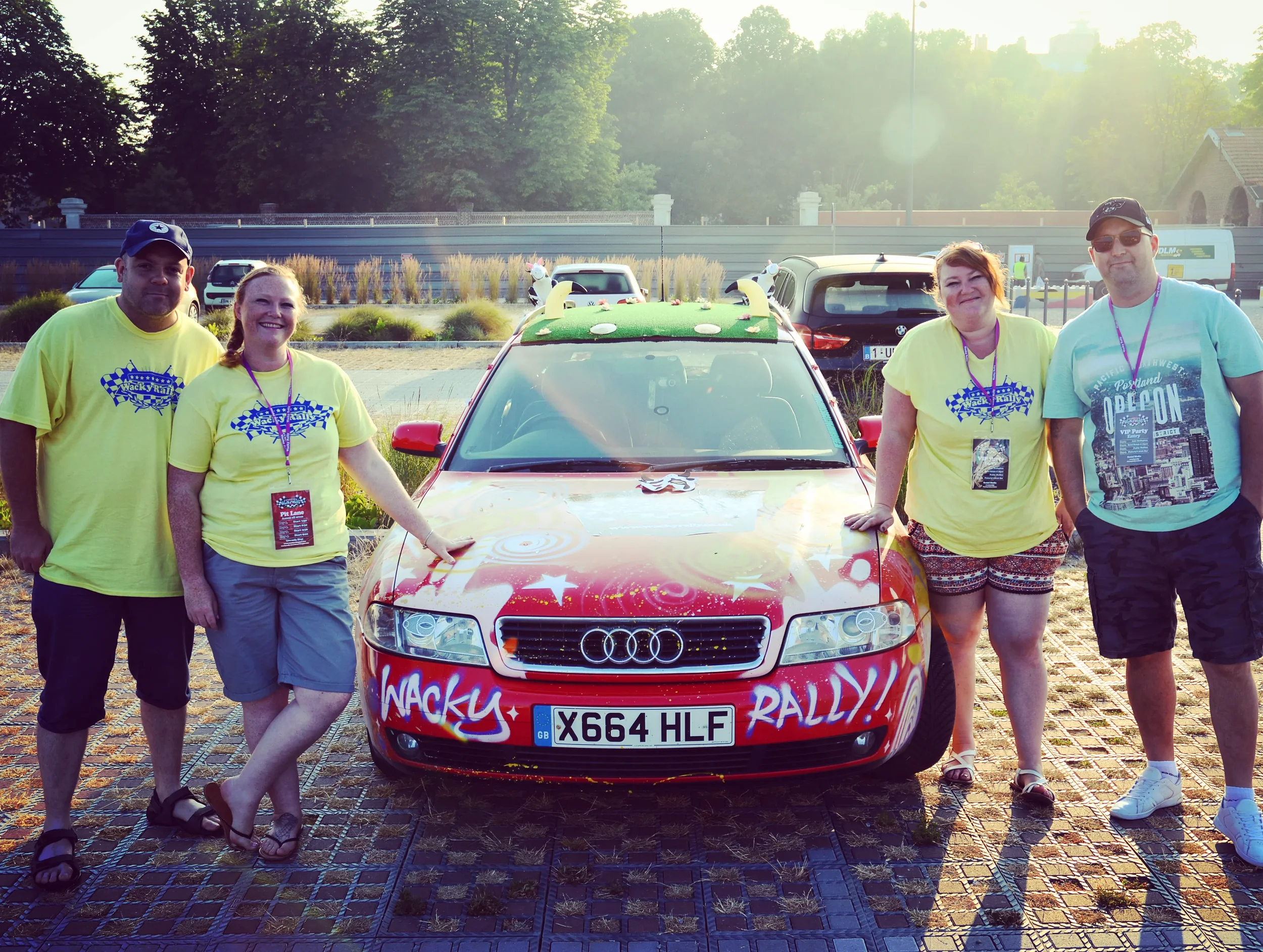 Four people standing next to a decorated red Audi car with rally-themed artwork. Two are wearing yellow t-shirts with a checkered flag logo, and two are in casual clothing. The scene is outdoors during sunset with trees and parked cars in the backgro