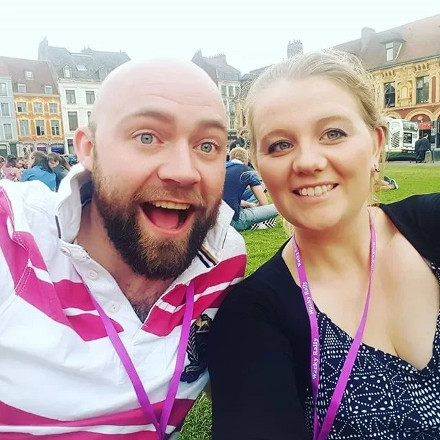 A man and woman taking a selfie outdoors in a grassy area with historic buildings in the background. The man has a beard and is smiling widely, wearing a white and pink striped shirt. The woman has blonde hair, is smiling, and wearing a black cardiga