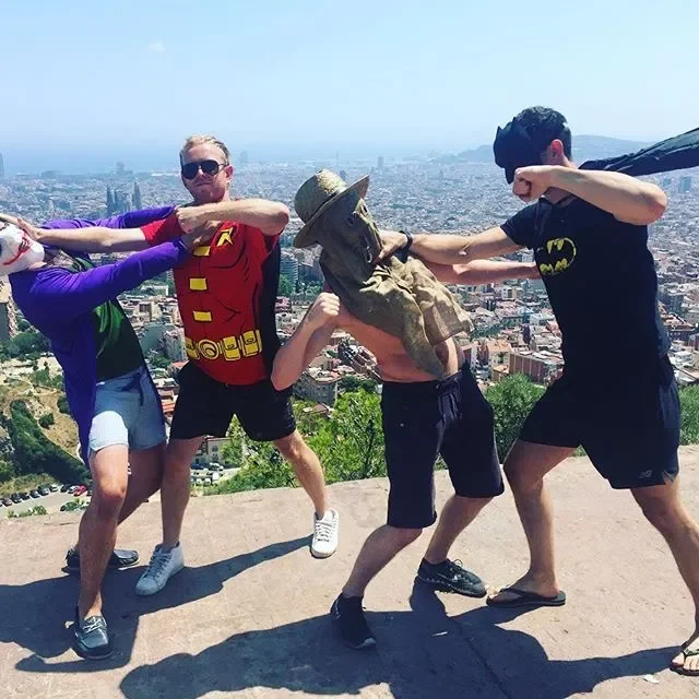 Four friends in superhero costumes playfully fighting with a large bronze bust sculpture of a man with a hat at a scenic overlook with city view.