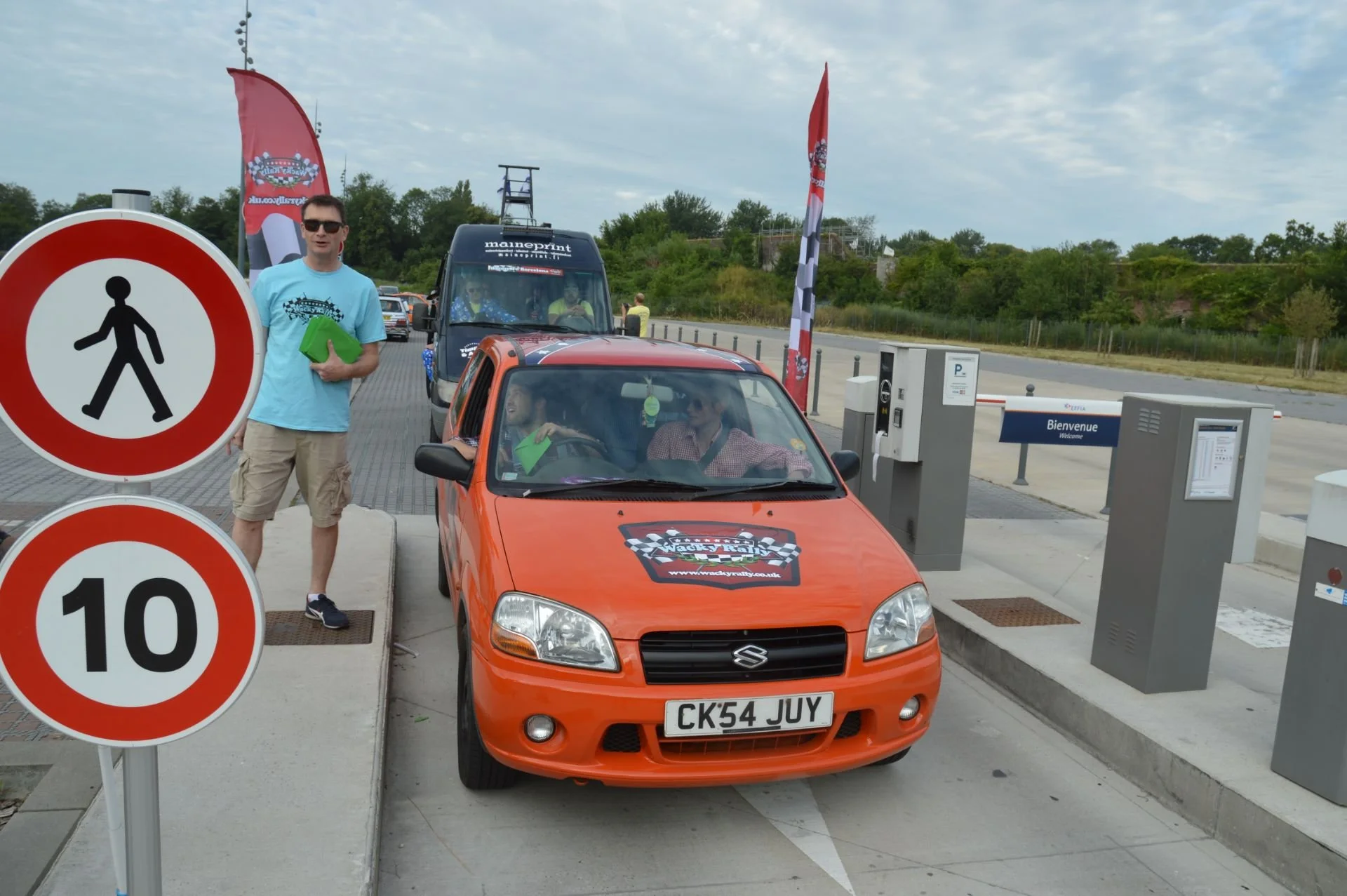 An orange Suzuki car with a race sticker on its hood, parked in front of parking machines at a car event. A man in a light blue shirt and shorts stands beside the car, holding a green booklet. Two women inside the car are talking, one holding a green