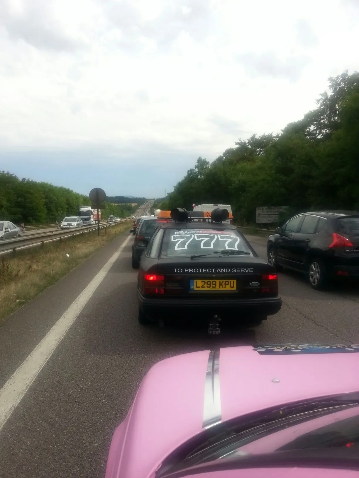 Traffic jam on a highway with police vehicle in front, decorated with the number 777 and the phrase 'To Protect and Serve' on the back, with other cars ahead and on the sides, and a cloudy sky above.