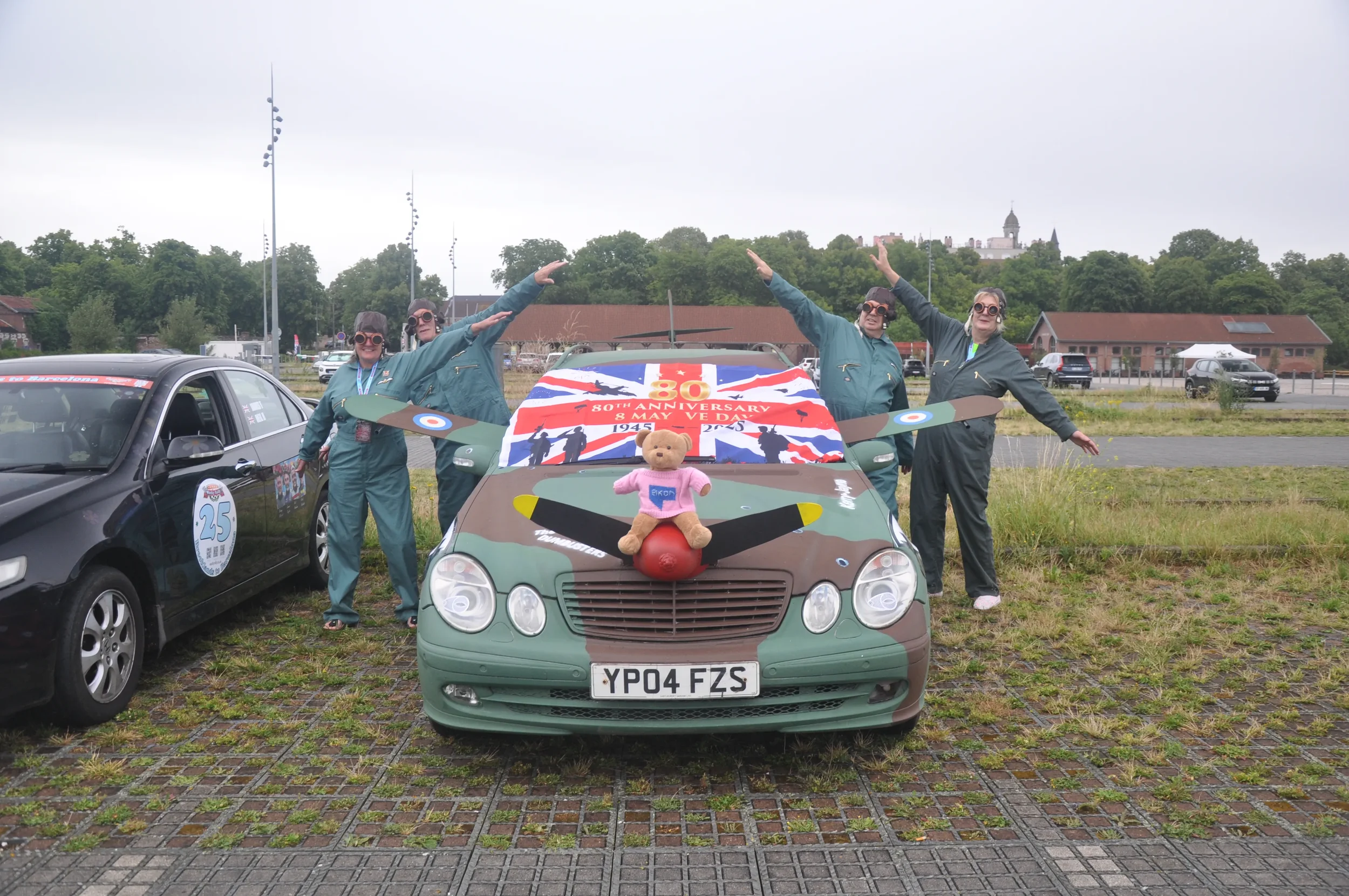 Four people dressed in aviation suits celebrate next to a decorated car with a teddy bear on the hood, during a 30th-anniversary event, with a banner in the background and parking lot in the distance.