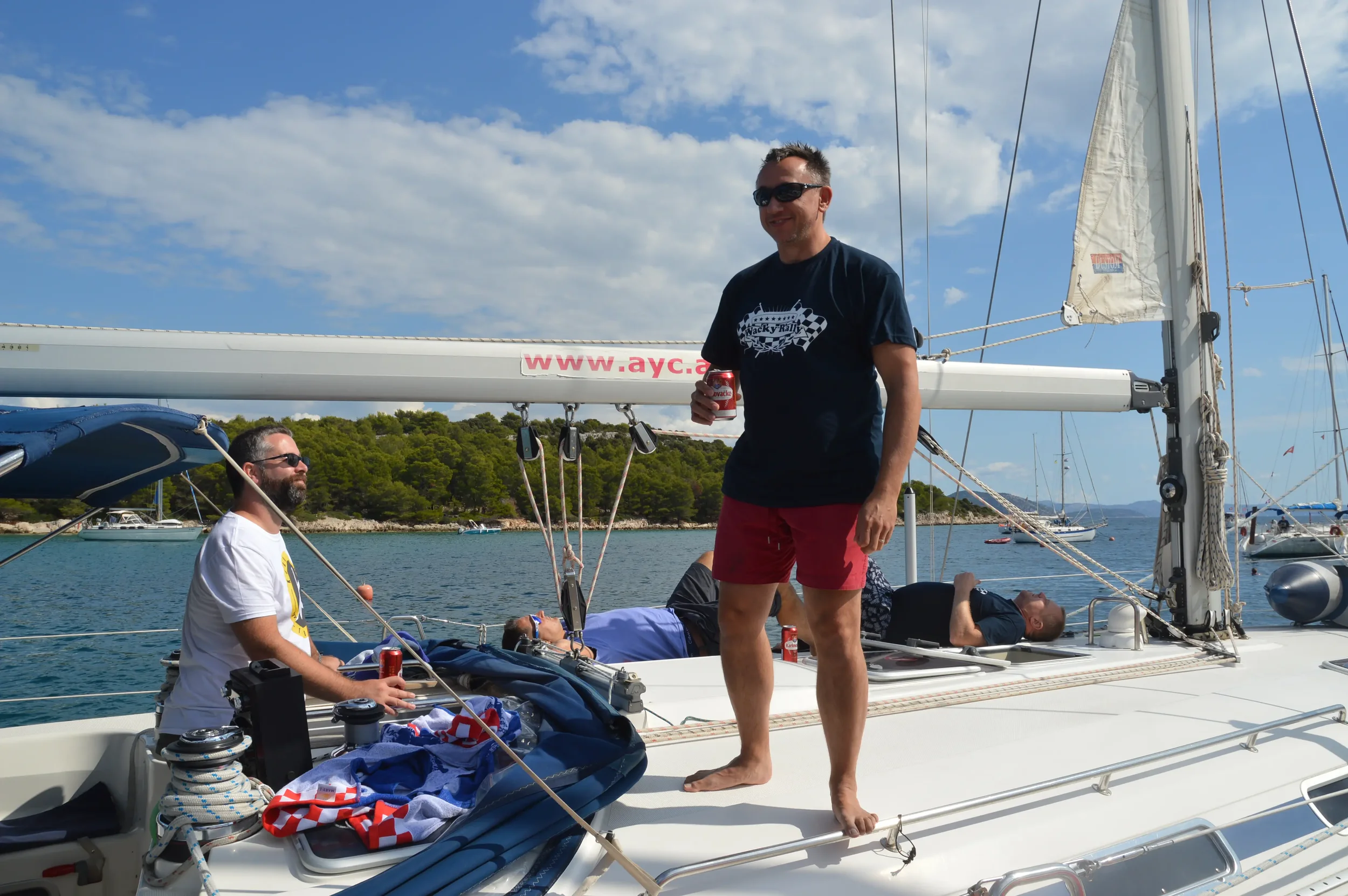 Four men relaxing on a sailboat with some drinking soda, one standing and three lying or sitting, in a peaceful water body with boats and green trees in the background under a partly cloudy sky.
