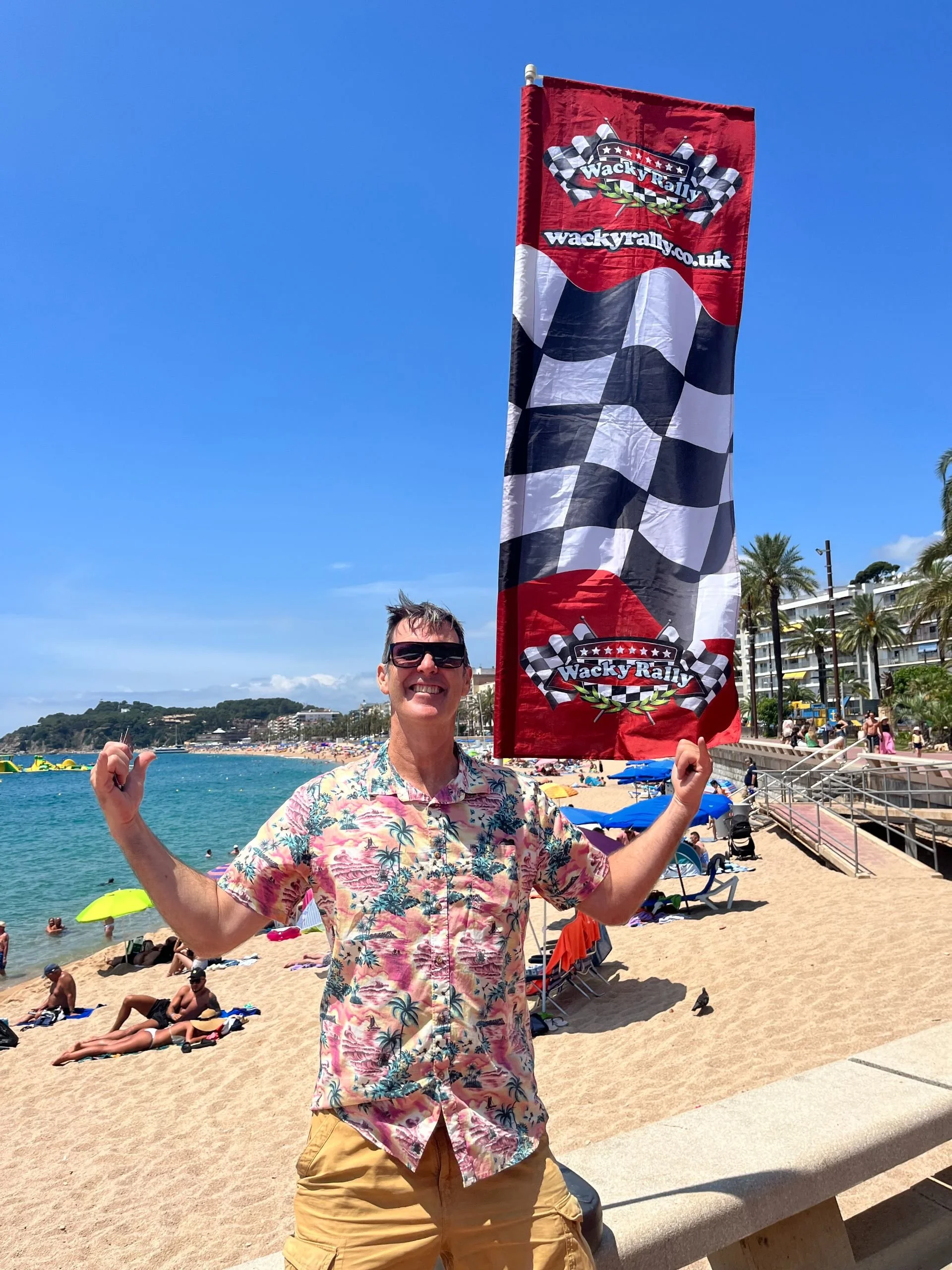 Man in sunglasses smiling and holding a Wacky Rally flag on a crowded beach.