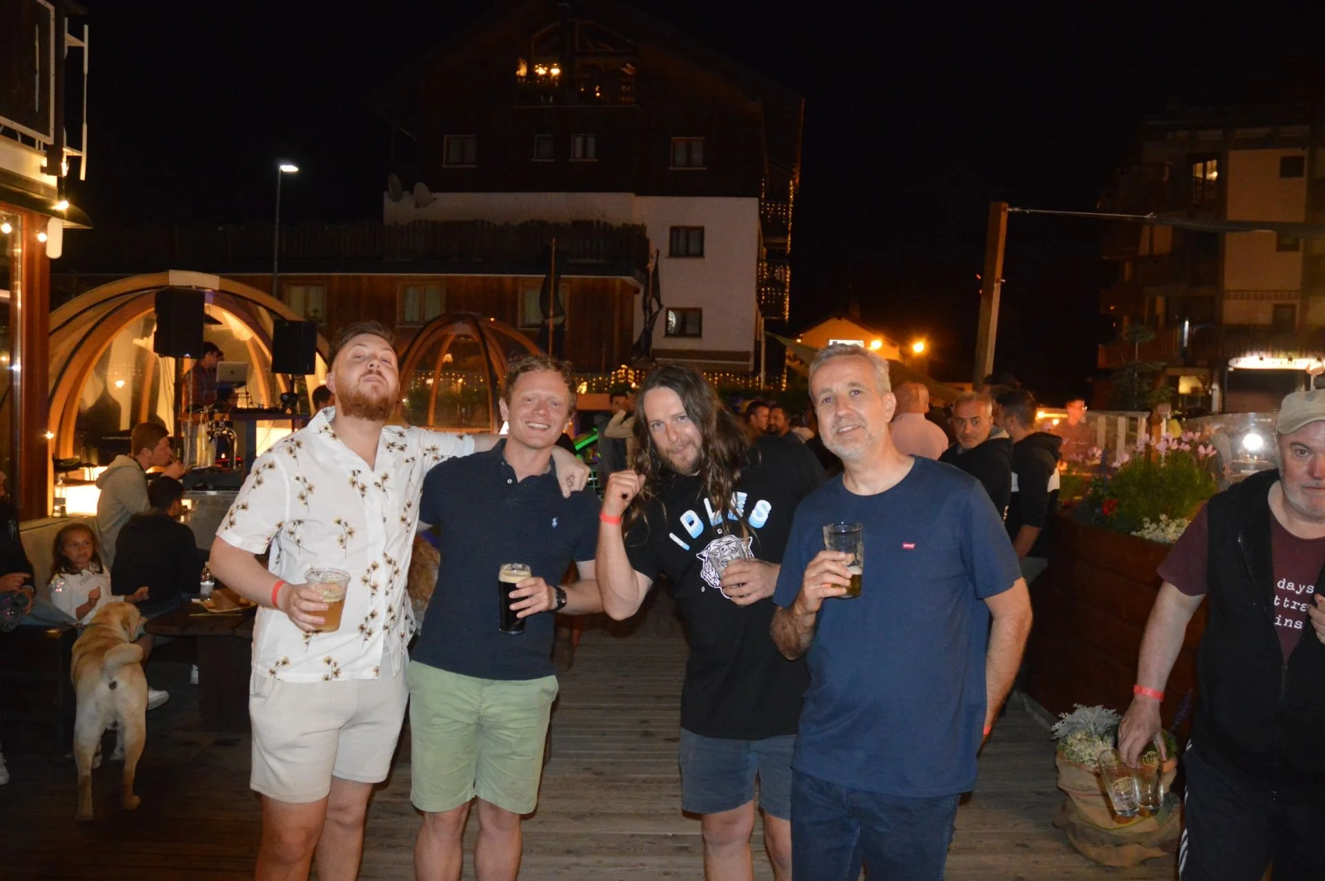 Four men standing together at an outdoor nighttime gathering, holding drinks, in front of a crowd of people and a bar with a wooden deck and decorative lighting.