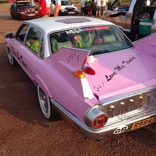 A customized vintage car painted pink and white with musical notes and the phrase 'Love Me Tender' on the hood. It has extra features like an angel wing and tail on the front, headlight covers, and a sticker on the windshield that says 'Barcelona.' T