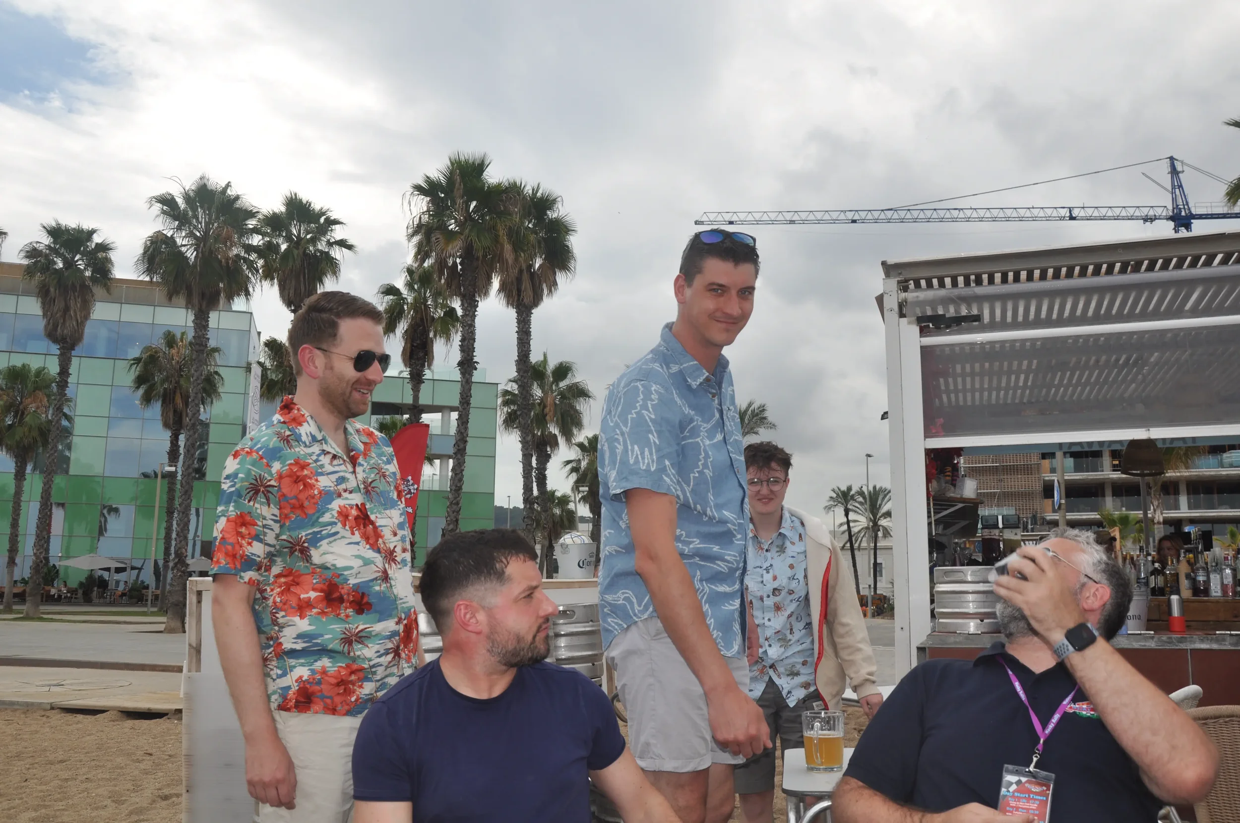 Group of five men socializing outdoors at a beachside bar with palm trees, modern buildings, and overcast sky in the background. One man is sitting, holding a beer, while four others stand nearby, one with a drink in hand.