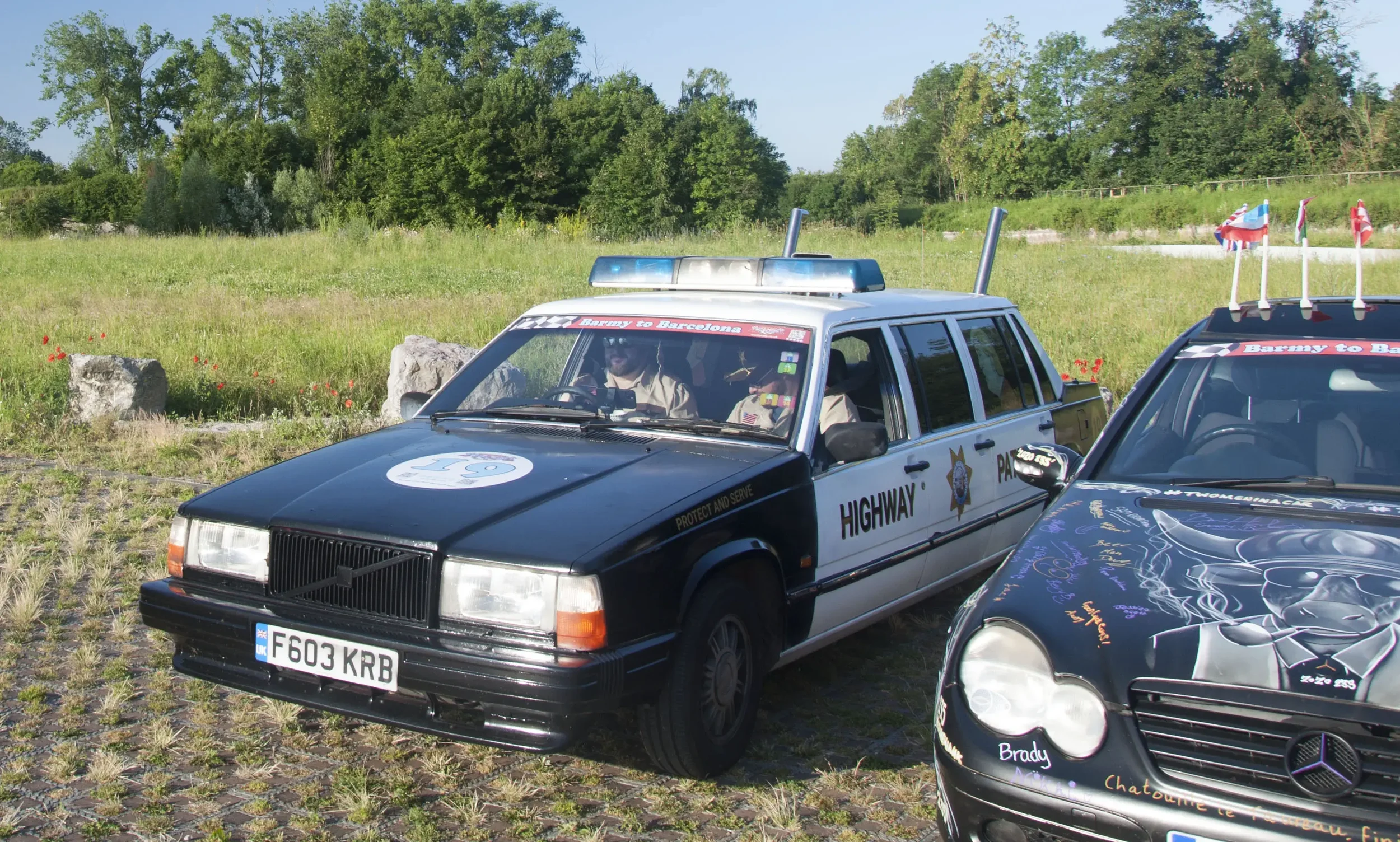 Two classic cars parked on a grassy field with trees in the background, one resembling a police vehicle and the other decorated with graffiti and stickers.