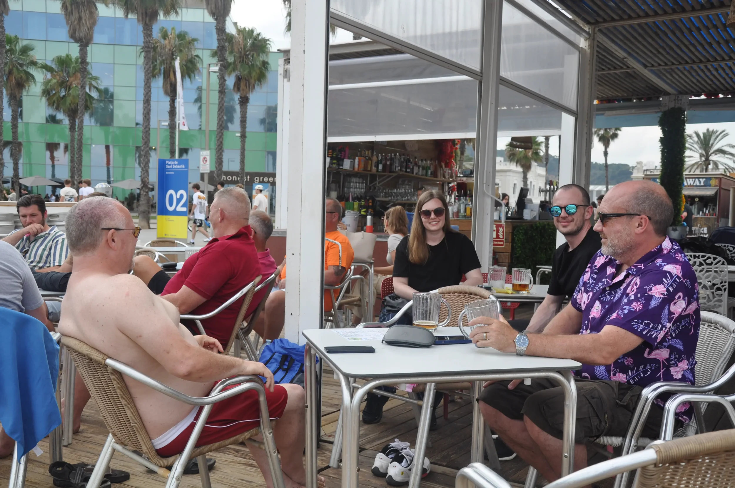 People sitting at outdoor tables of a bar or restaurant near palm trees, enjoying drinks and socializing.