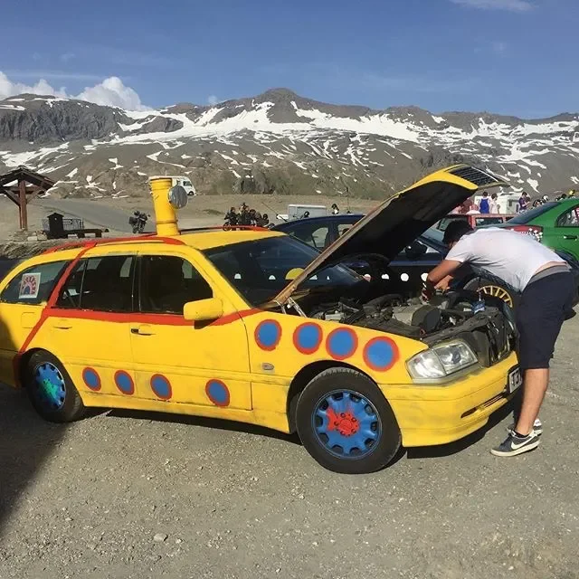 A yellow car with colorful circle and dot designs on it, parked outdoors with the hood open. A man is working on the engine. In the background, there are other cars, a yellow pipe, people, and snow-capped mountains under a blue sky.
