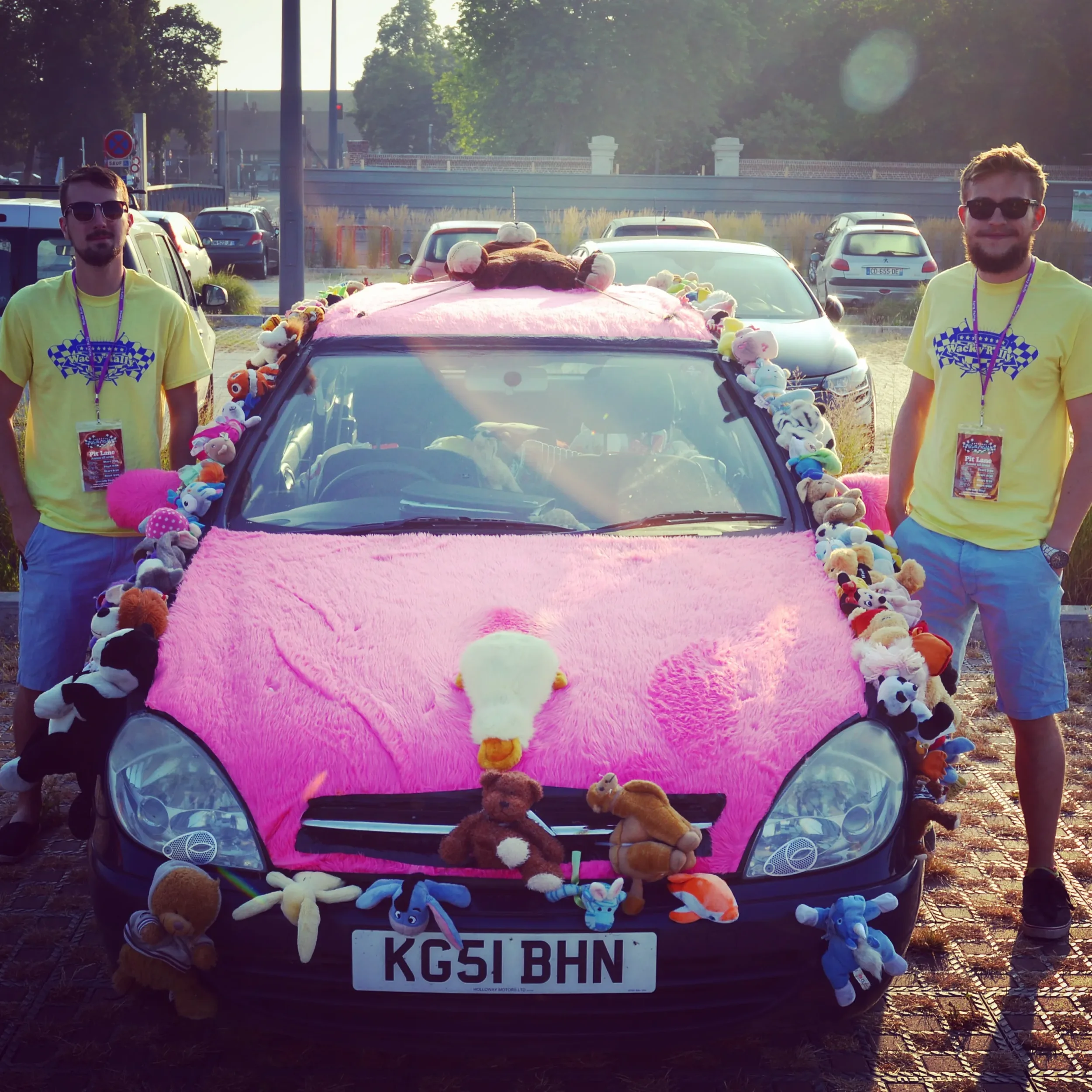 A black car decorated with pink fluffy material on its hood and roof, adorned with numerous stuffed animals including bears, pandas, and other plush toys. Two men wearing yellow t-shirts and sunglasses are standing on either side of the car. The back