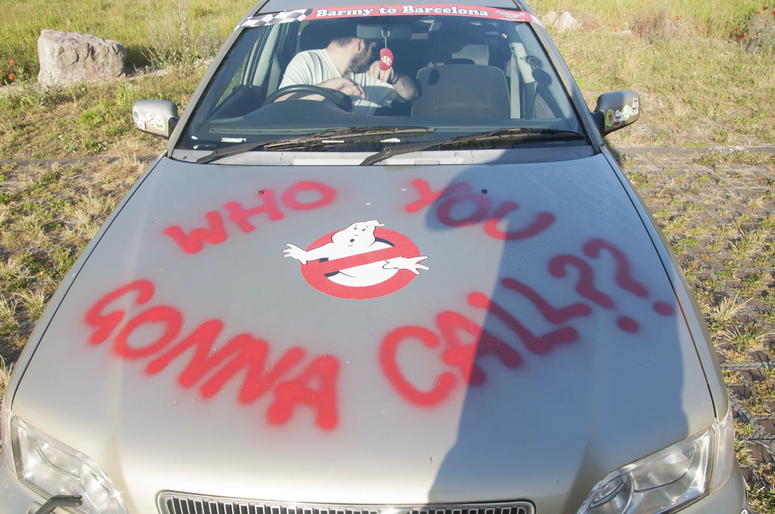 A silver car parked outdoors with a Ghostbusters logo and the phrase 'Who you gonna call??' spray painted on the hood. The driver is a man looking down and adjusting something inside the car.