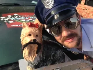 A man dressed as a police officer taking a selfie with a small dog wearing sunglasses and a red bow on its head.