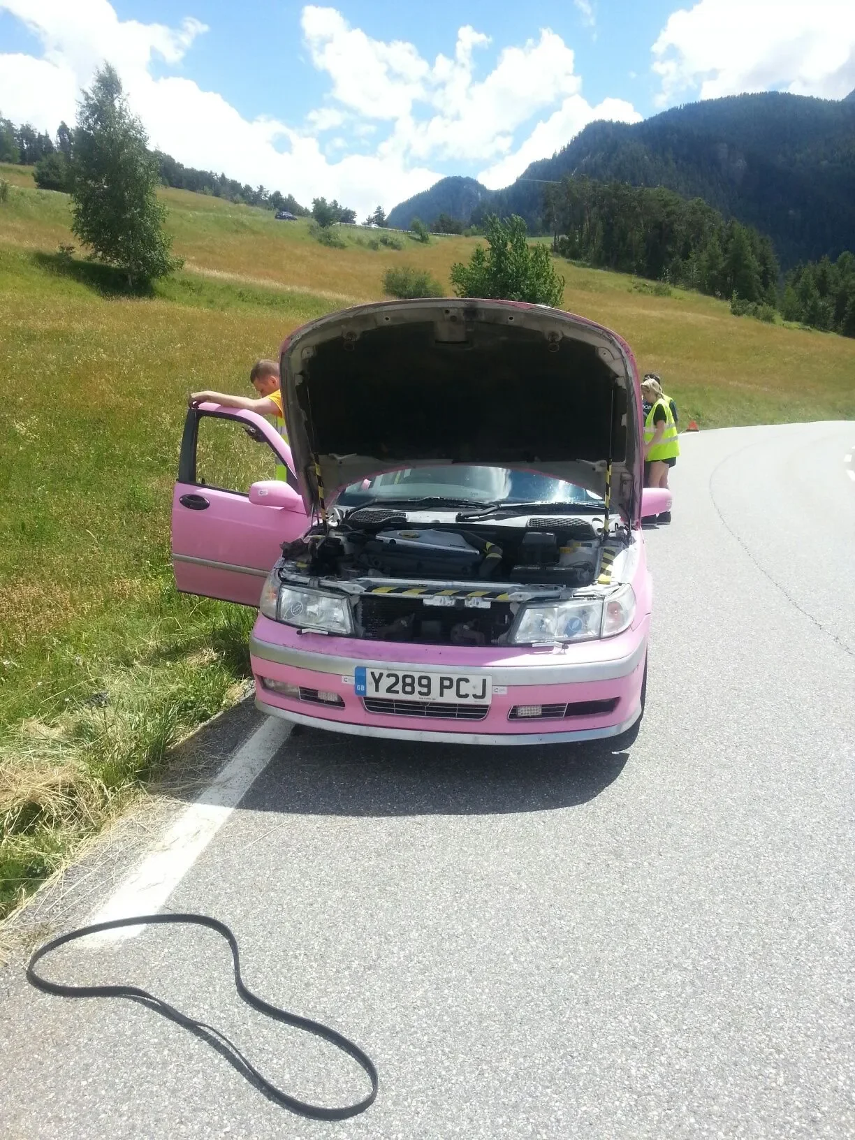 Pink car parked on the side of a highway with its hood open and the driver’s side door open, roadside assistance or a mechanic inspecting the engine, rural scenic background with green hills, mountains, and cloudy sky, and a black cable on the road.