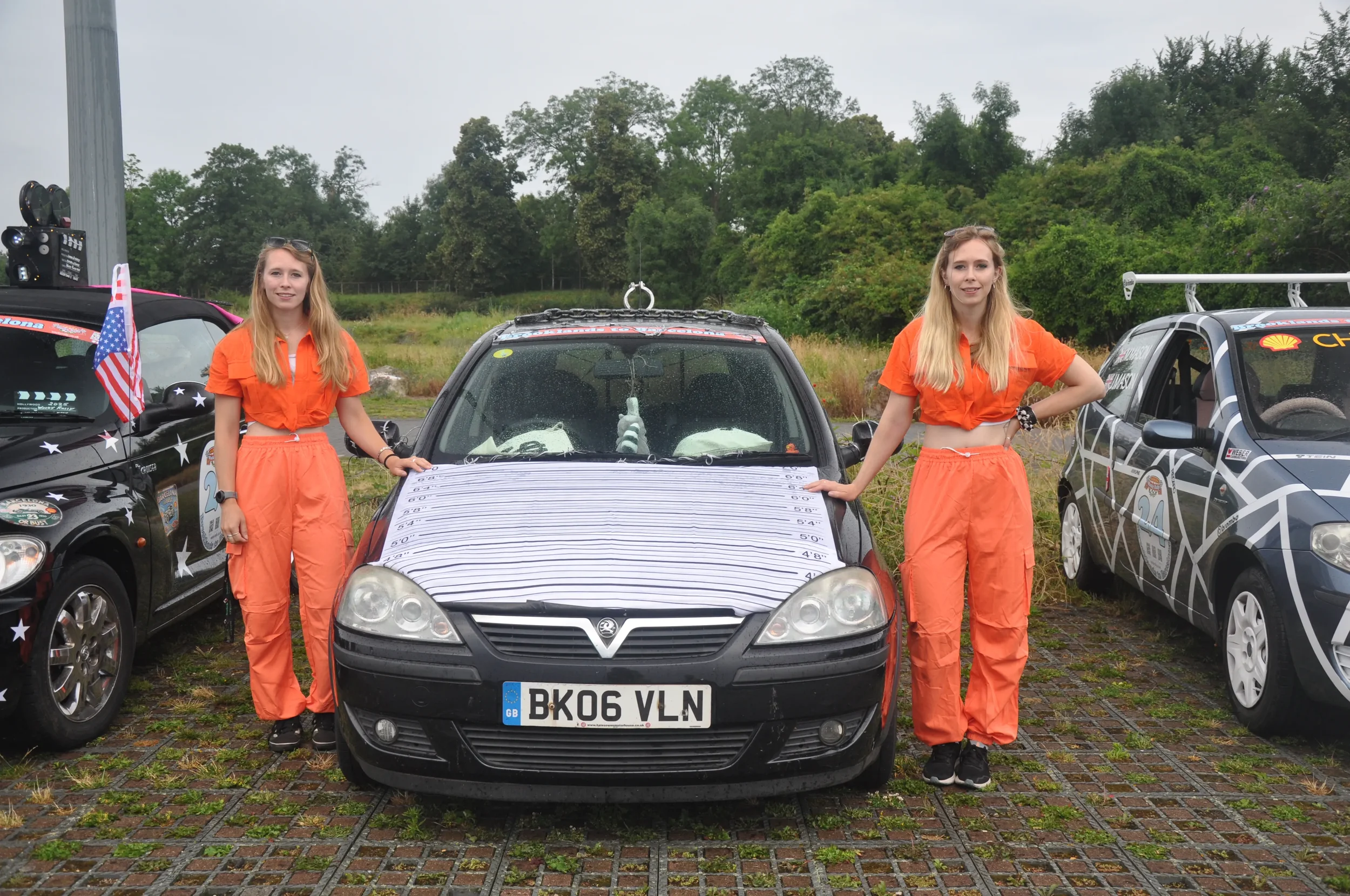 Two young women dressed in orange jumpsuits stand beside a black car with a height measurement chart on the hood, in a parking lot with other decorated cars and green trees in the background.