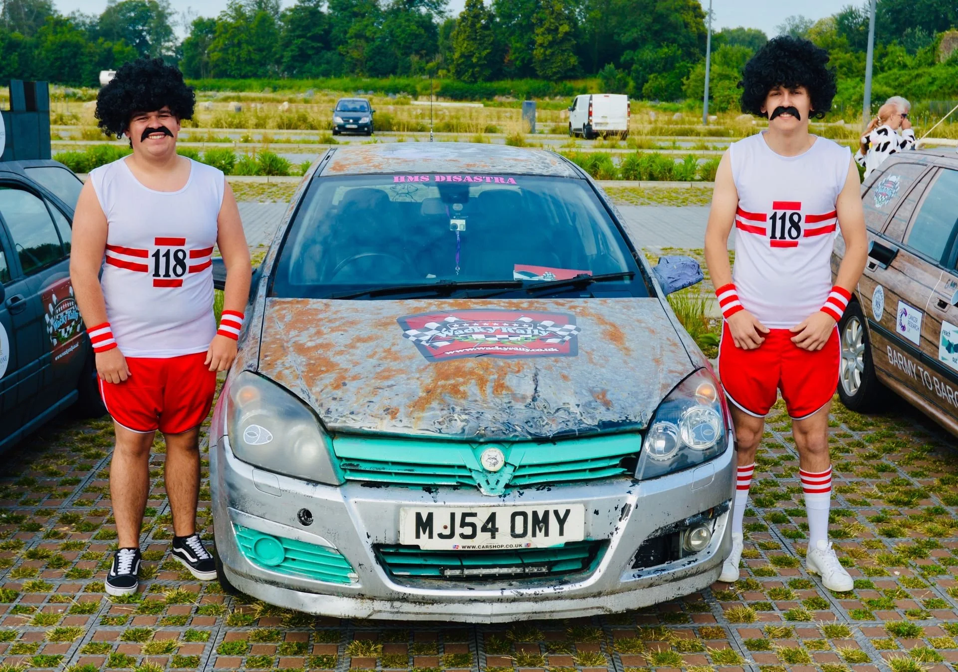 Two people dressed as sumo wrestlers in old, rusty racing suits with curly black wigs and mustaches, standing on either side of a rusty, weathered car with racing stripes and a sign on the windshield that reads "HMS Disastra." The car has a customize