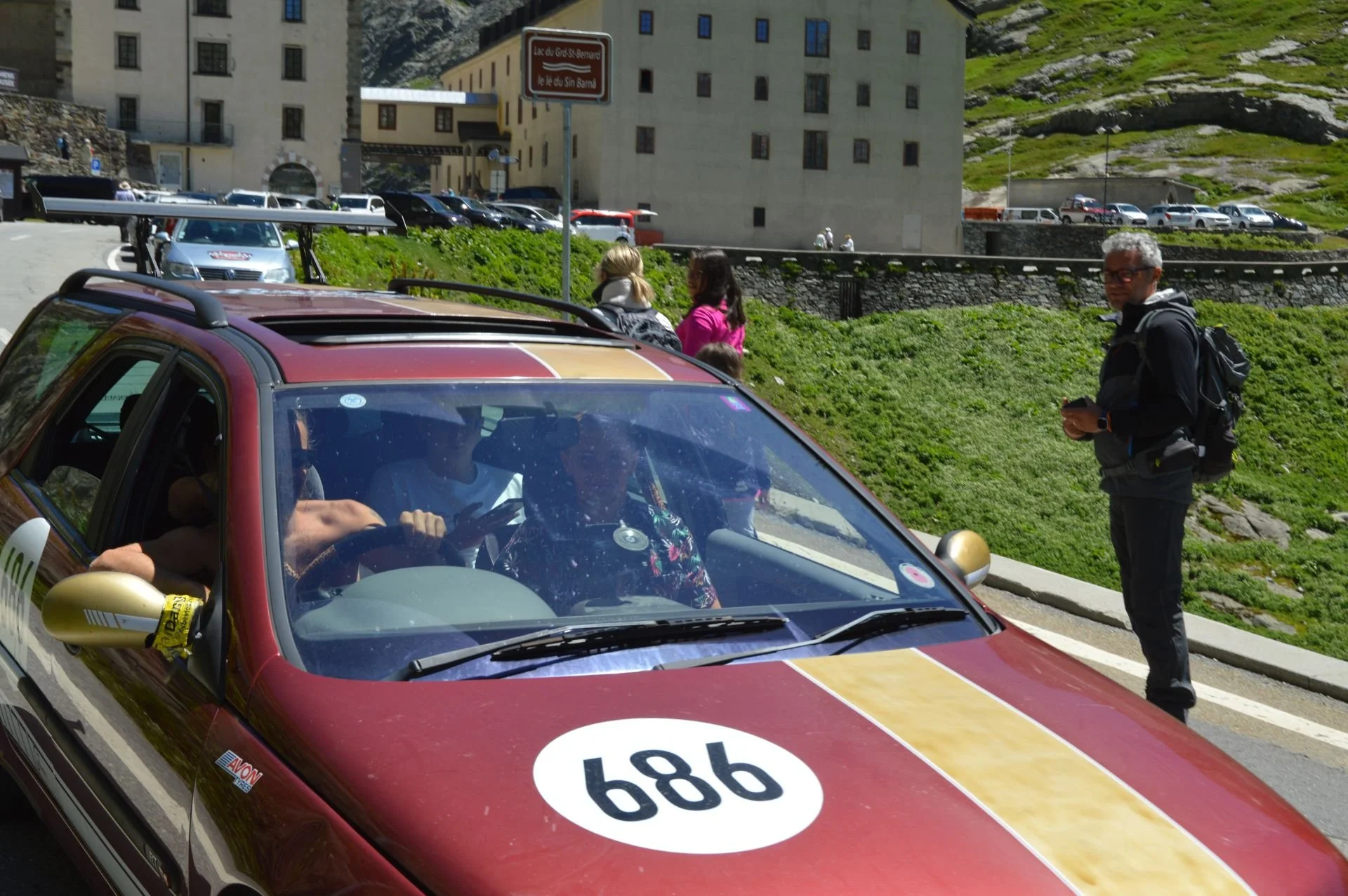 A vintage racing car with number 986 on its hood parked on the street, with people standing nearby and a person inside the car. There is a man with gray hair and glasses standing next to the car, holding a phone. The background features a building, s