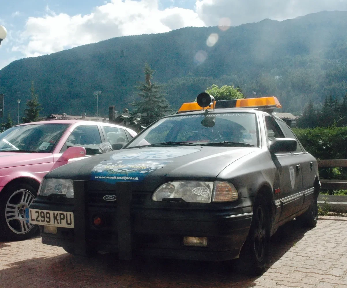 A black Ford police car with a yellow light on top and a loudspeaker, parked next to a pink car in an outdoor lot with mountains and cloudy sky in the background.
