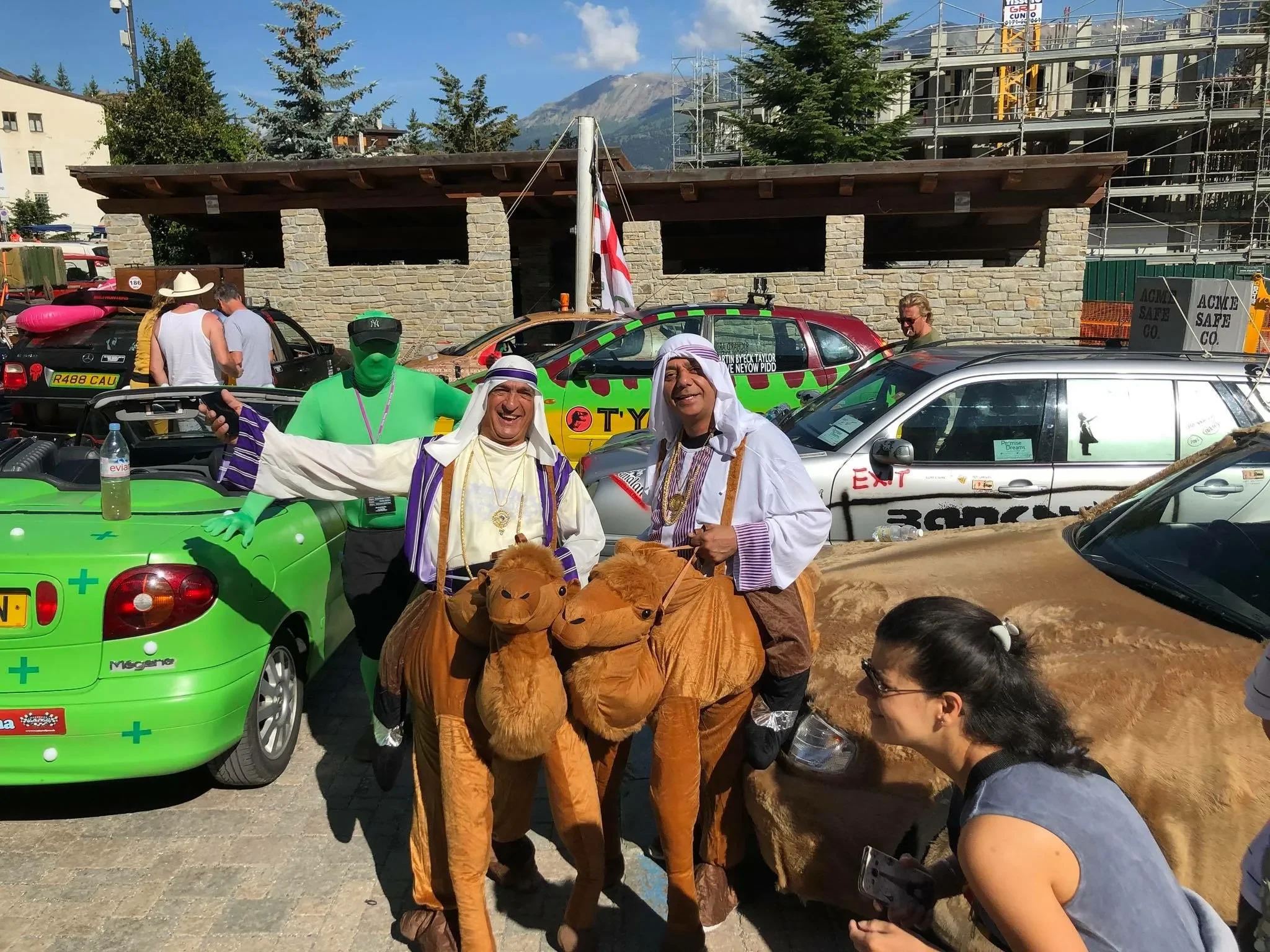 People in costumes, including individuals riding on inflatable camel costumes and others dressed as Middle Eastern characters, are gathered in an outdoor setting with parked cars and a stone building in the background. The scene appears festive with 