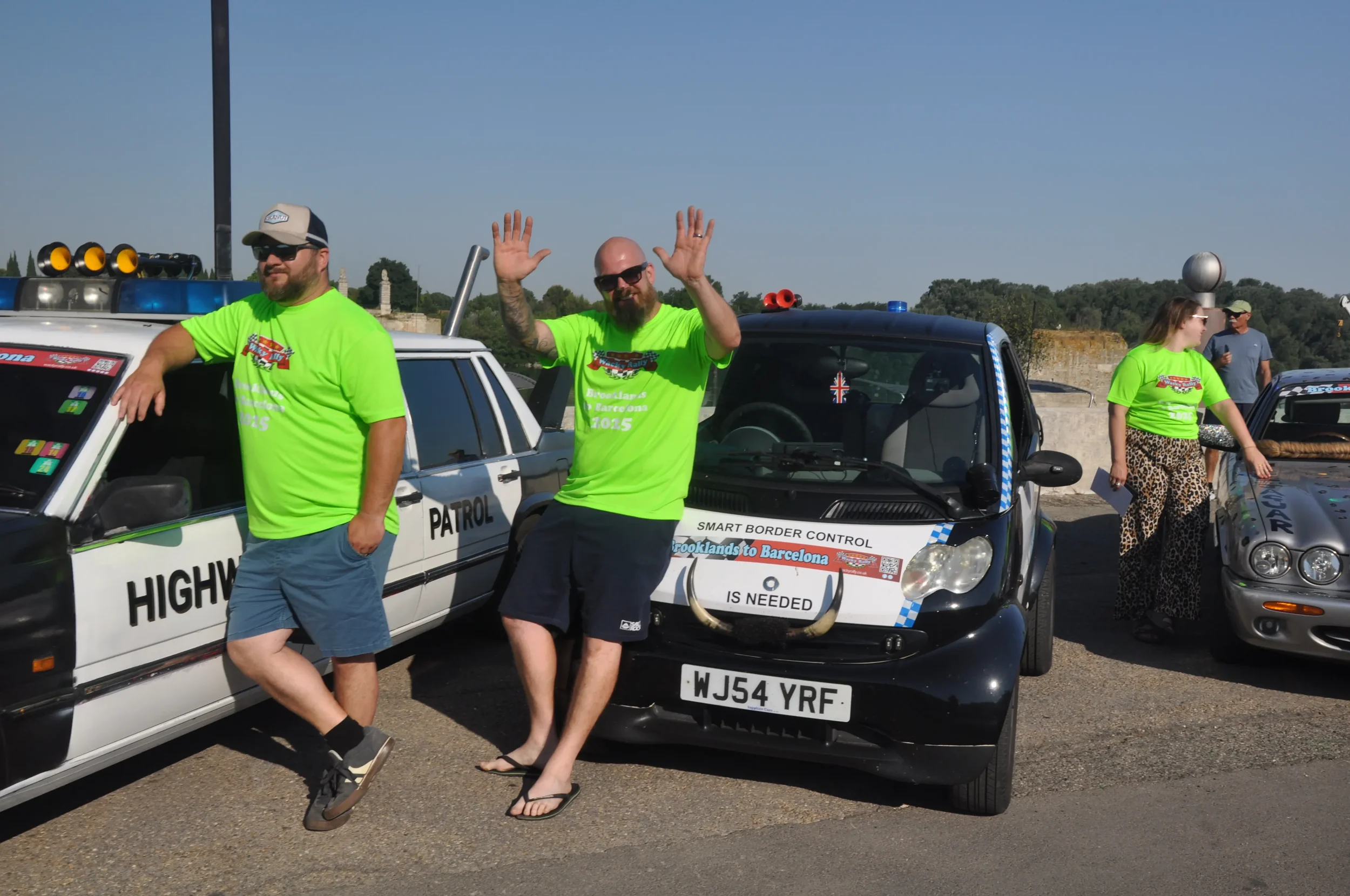 Three people in bright green shirts standing next to police-style cars at a car event, with two men posing and a woman gesturing towards a vehicle.