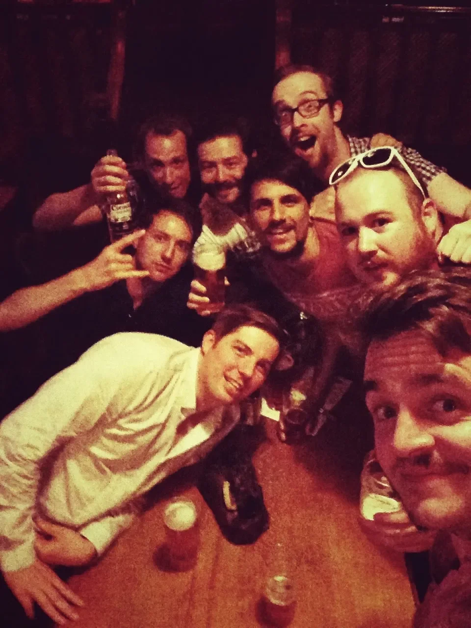 Group of friends having fun, posing for a selfie at a bar or party, holding drinks, smiling, and making playful gestures.