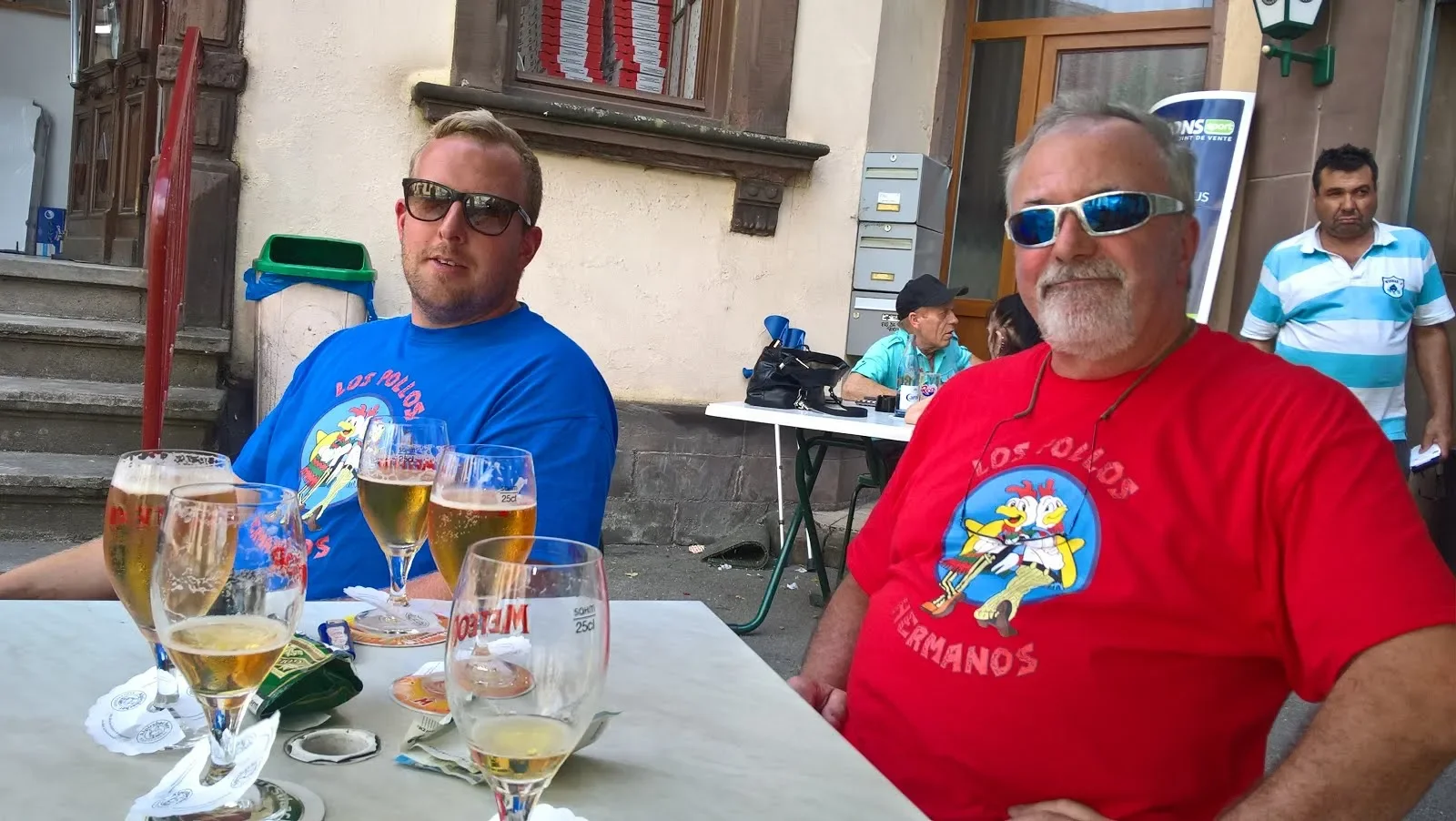 Two men sitting at an outdoor table, smiling, wearing sunglasses and colorful T-shirts with a rooster logo. Several glasses of beer are on the table. In the background, other people are sitting and walking near a building with stairs and mailboxes.