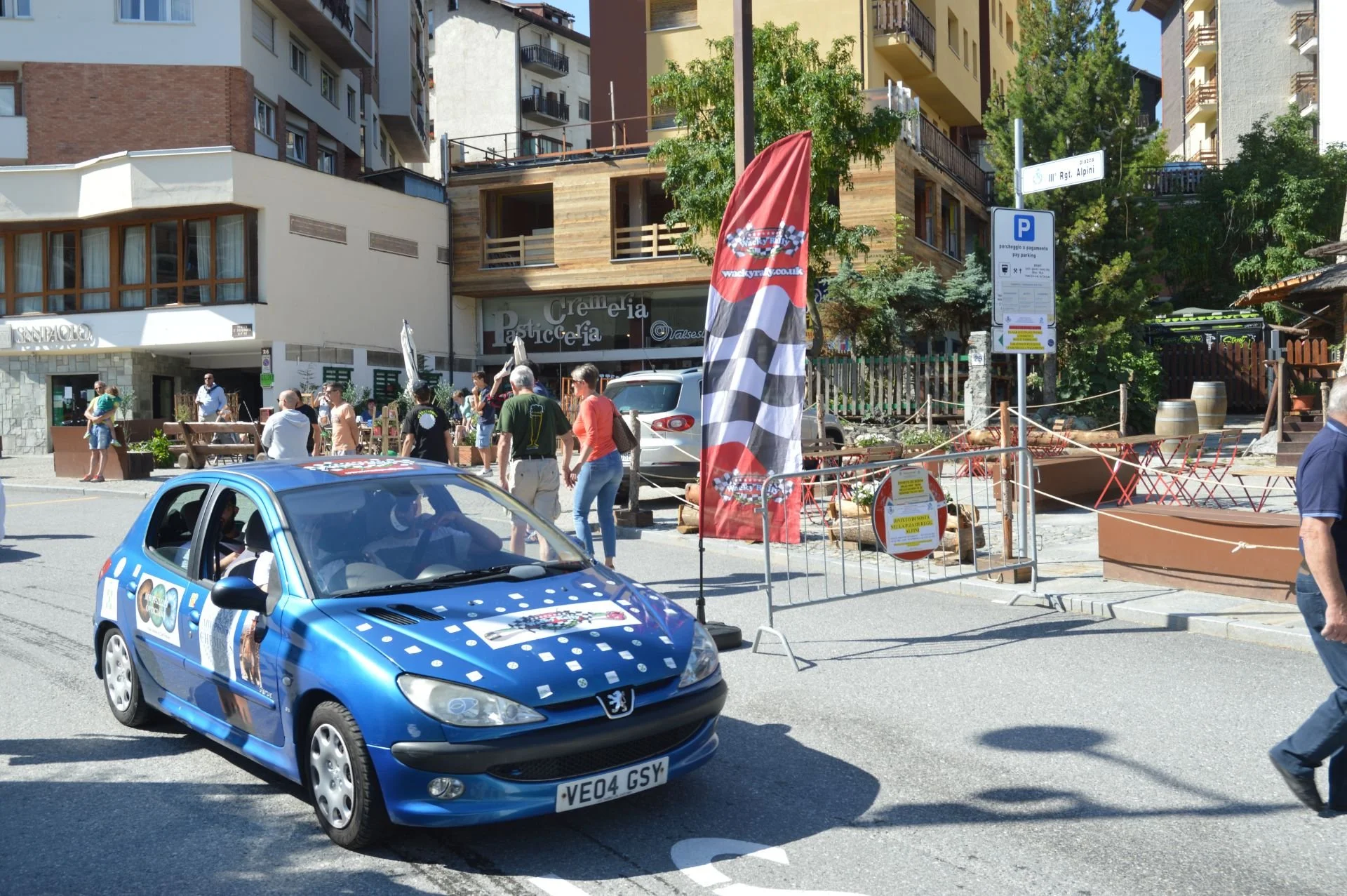 A street scene with a blue Peugeot car in the foreground and a group of pedestrians in the background near outdoor seating and banners, with multi-story buildings and trees surrounding the area.