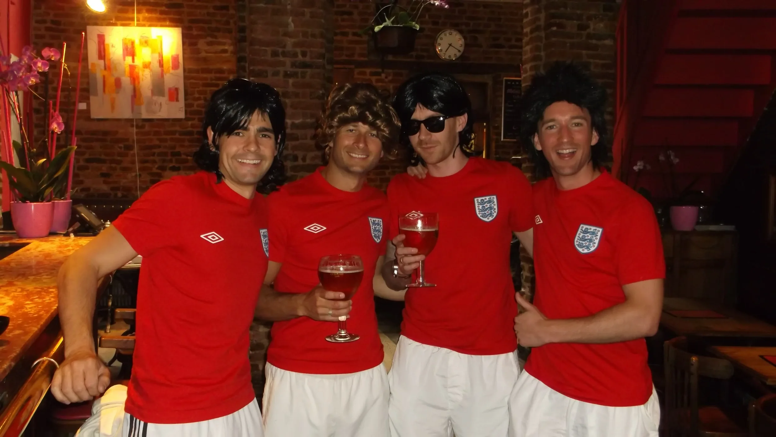 Four men dressed as soccer fans in red England jerseys and white shorts, holding glasses of beer, standing together in a bar or restaurant with brick walls and colorful artwork behind them.