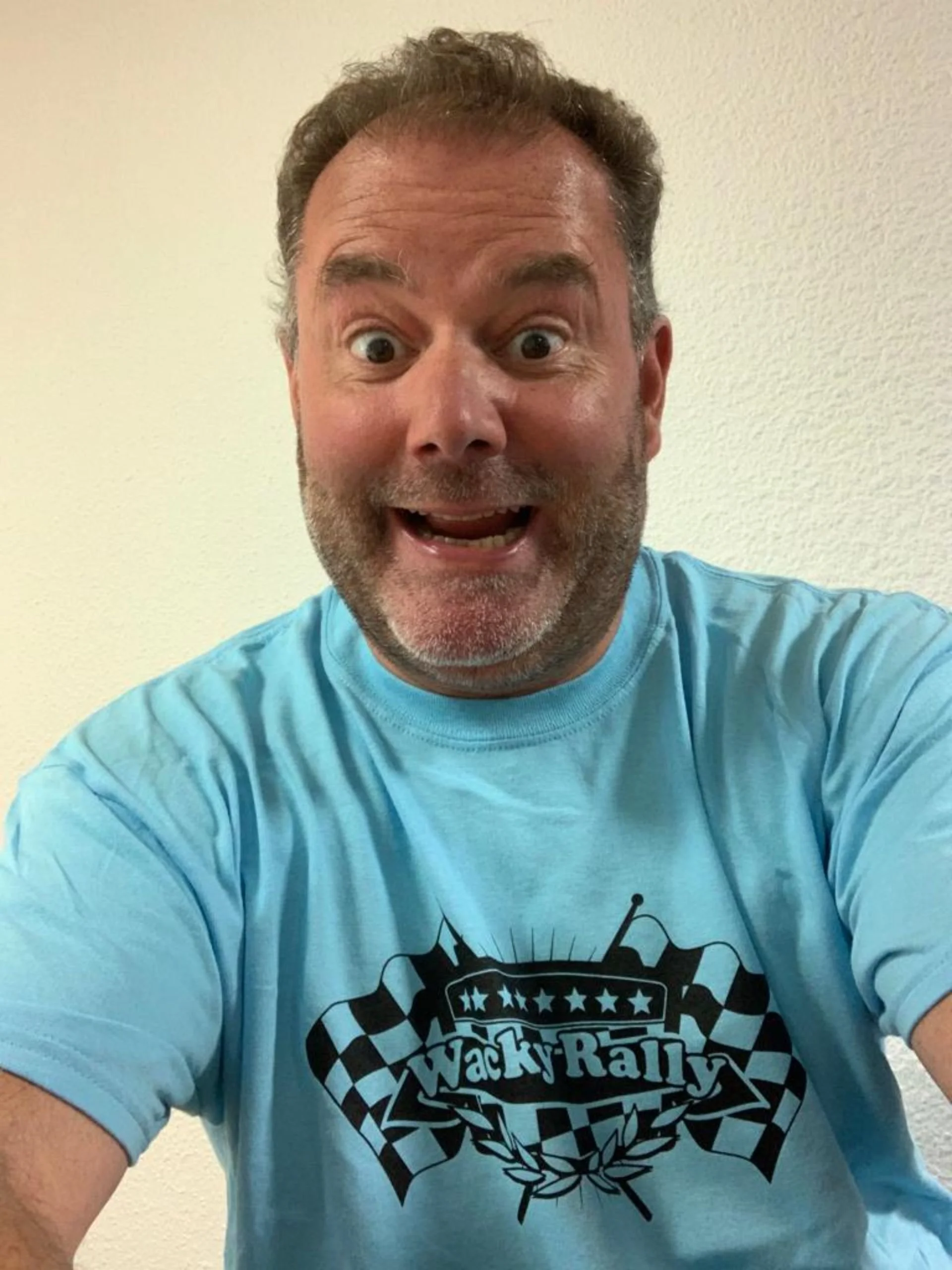 A man taking a selfie, wearing a light blue T-shirt with a racing-themed graphic and text that says 'Wacky Rally.' He has short, curly hair, a beard, and is smiling with an open mouth.