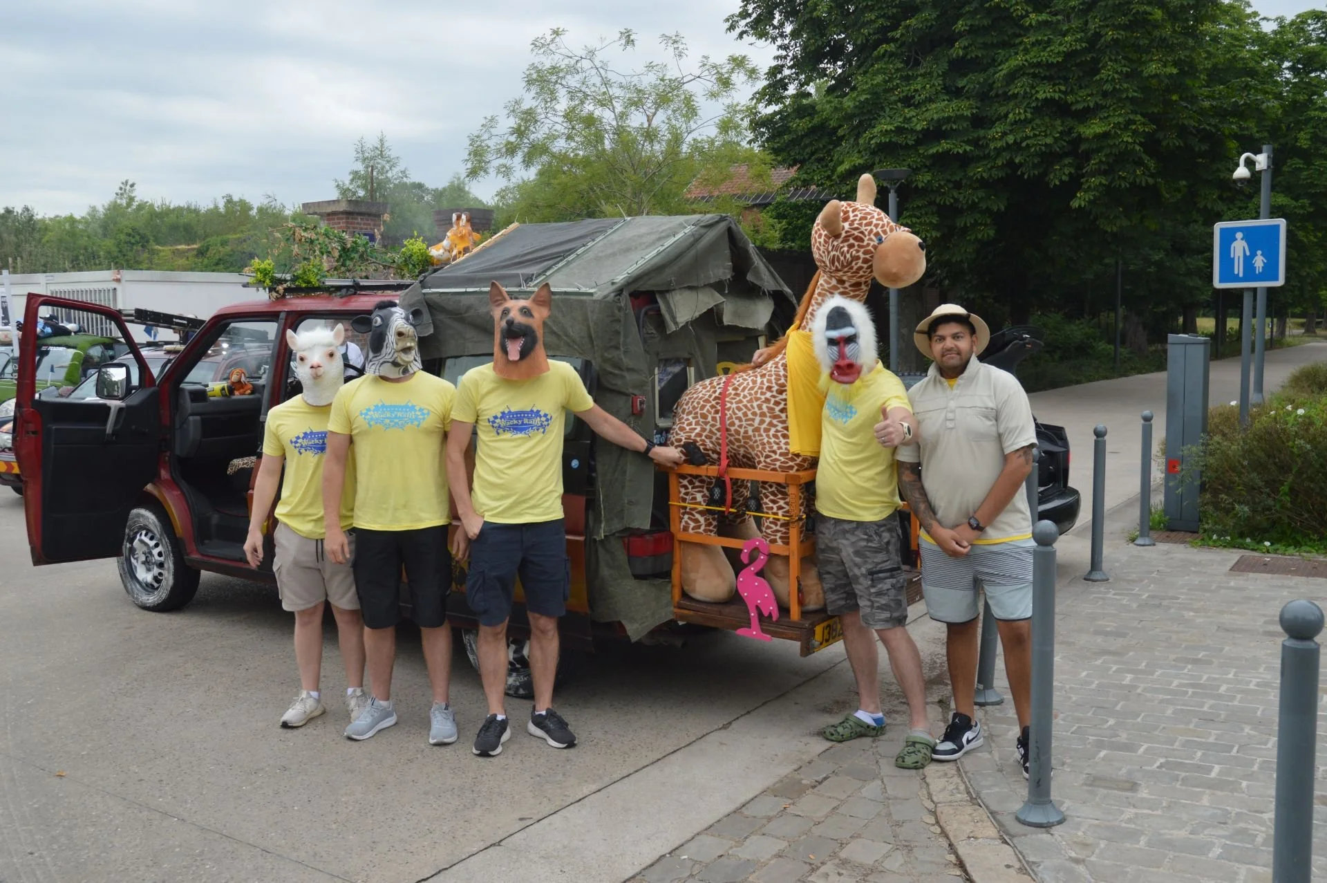 Group of people wearing animal masks, standing next to a decorated vehicle with a giraffe inflatable toy on the back, in an outdoor parking lot.