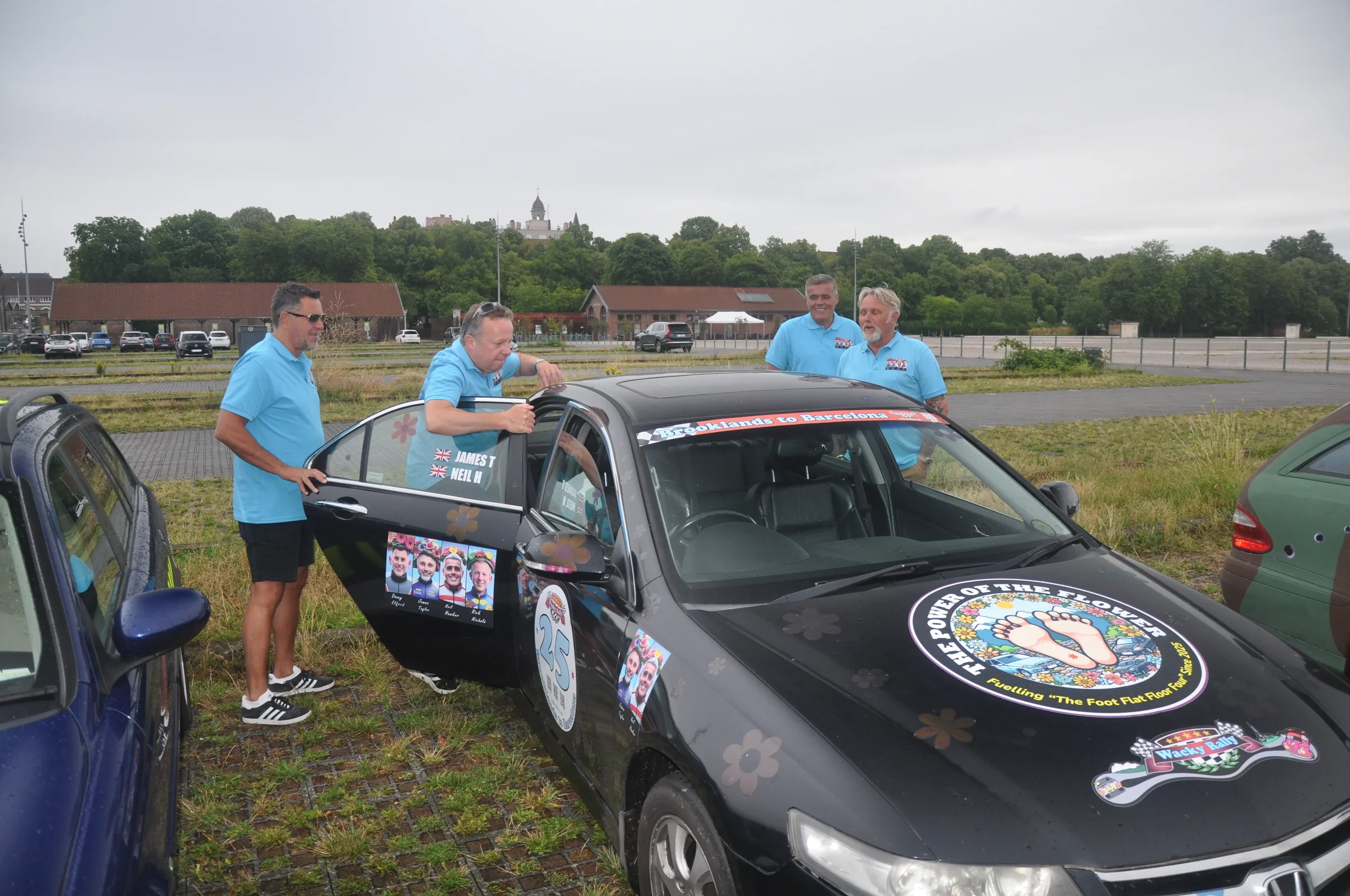 Four men in blue shirts standing around a black race car with logos and stickers, parked in a grassy area with other cars and trees in the background.