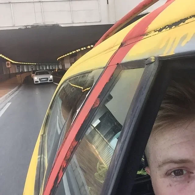 Close-up of a motorcyclist wearing a yellow and red helmet, with a car driving underneath a bridge in the background.