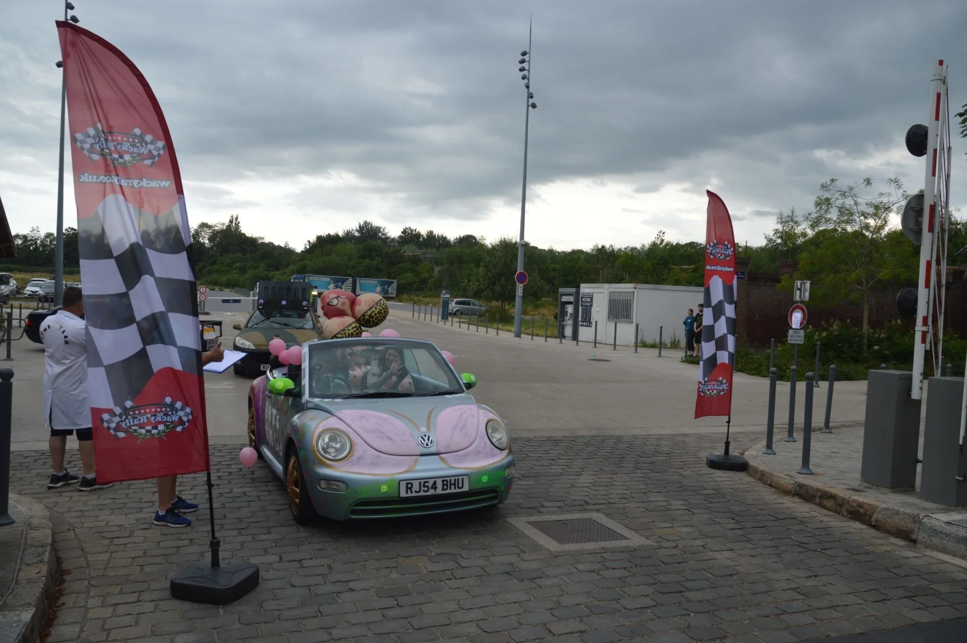 Colorful Volkswagen Beetle convertible decorated with large balloons, including a teddy bear, at a car event with Goodyear flags and a checkered racing pattern backdrop.
