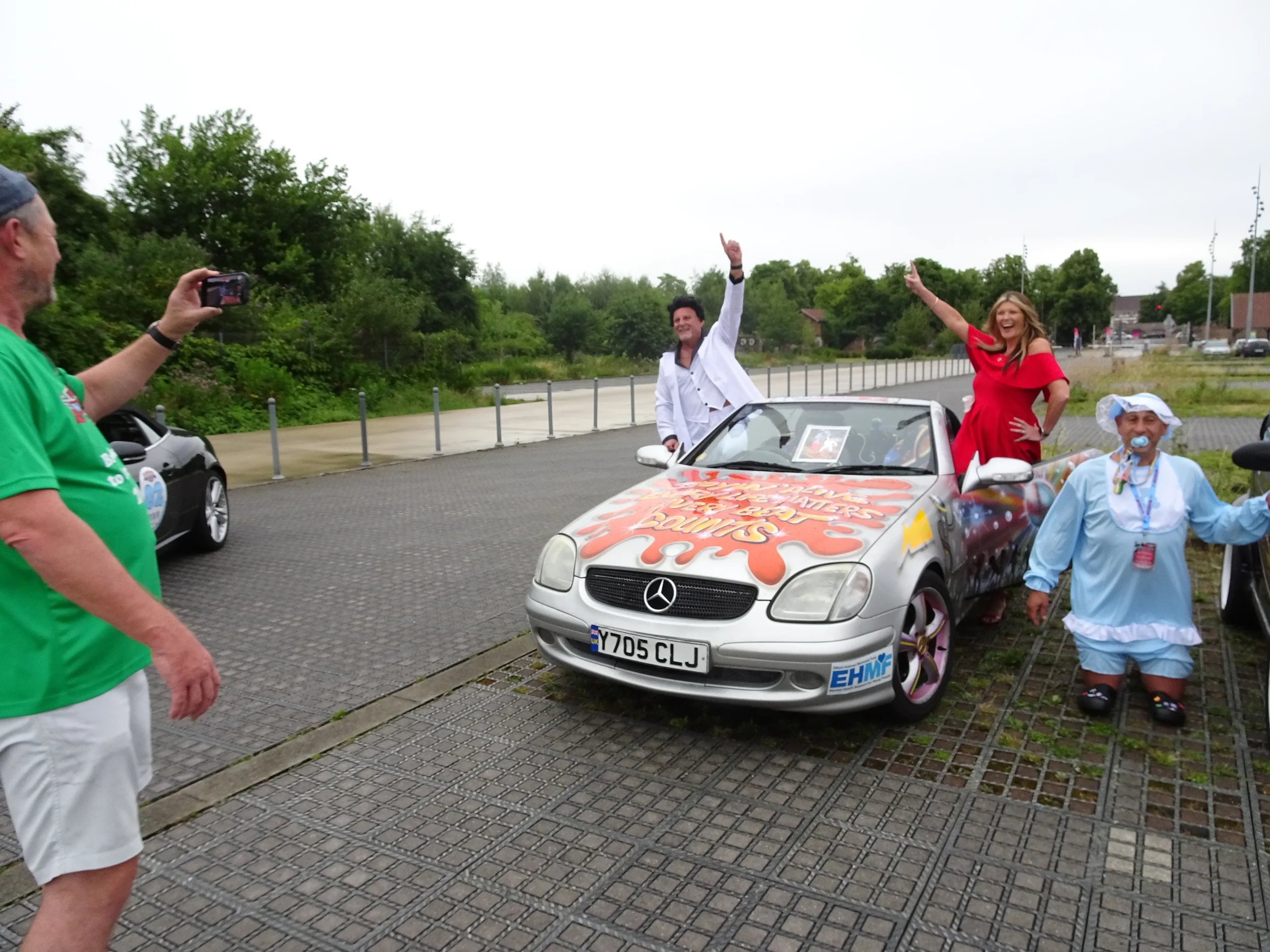 People celebrating around a decorated Mercedes Benz car, with two women in colorful dresses on the car and a man in a white jacket waving, while a man in green takes a photo, in an outdoor parking lot.