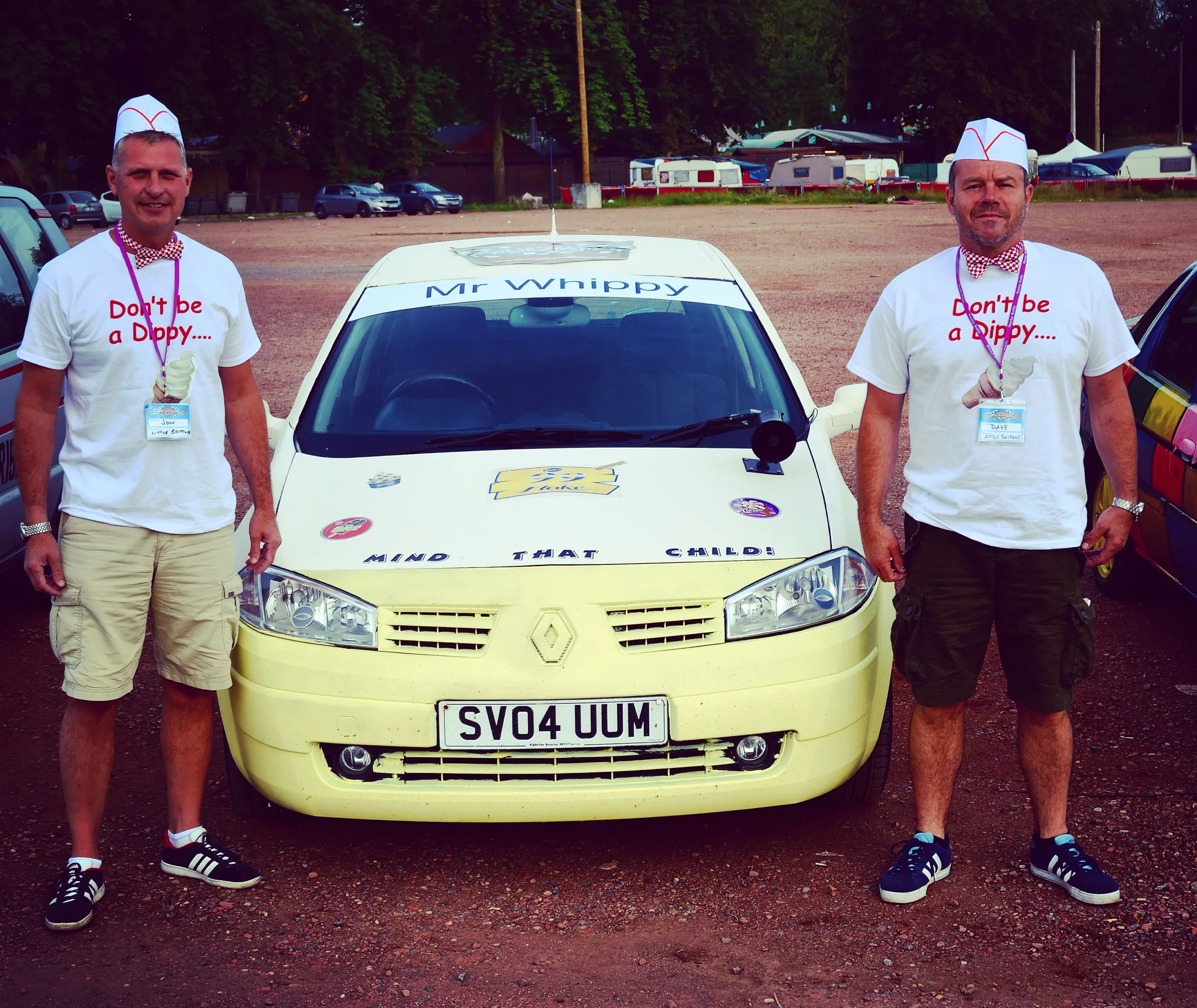 Two men standing next to a yellow Renault car with a sign on the roof that says 'Mr Whippy.' They are in a parking lot with other cars and trailers in the background. Both men are wearing white T-shirts with the slogan 'Don't be a Dippy...' and ice c