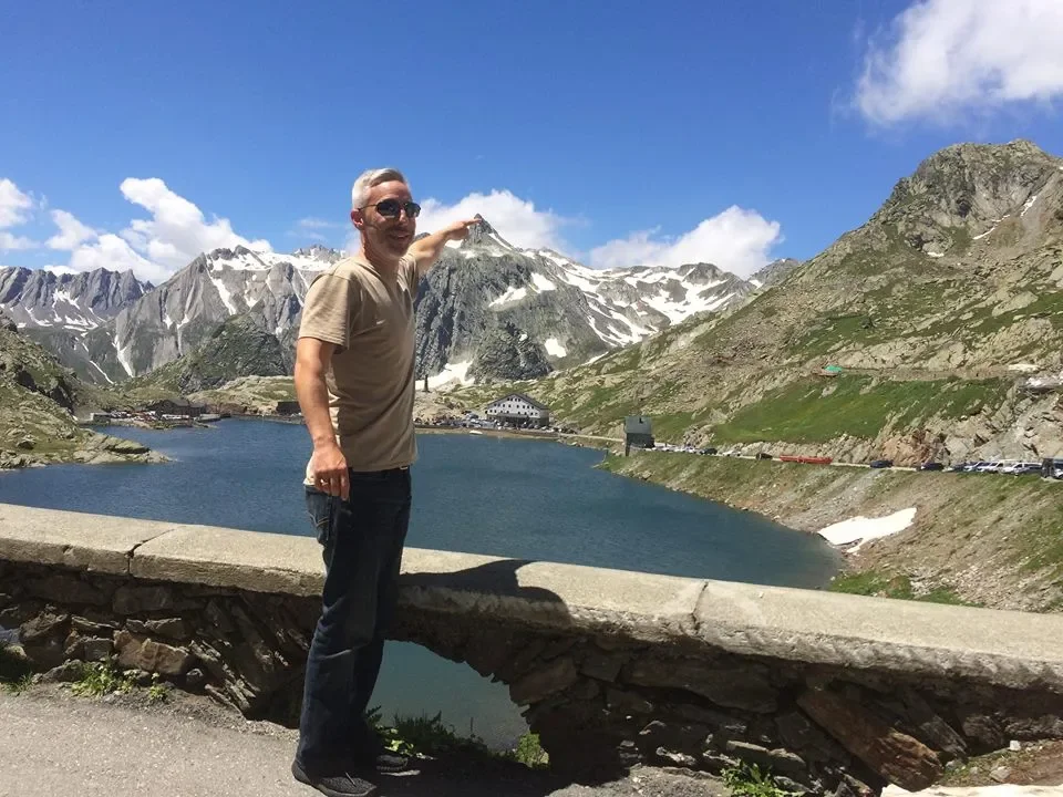 A man in sunglasses and a beige t-shirt pointing toward a snow-capped mountain in a scenic landscape with a lake and mountains in the background.