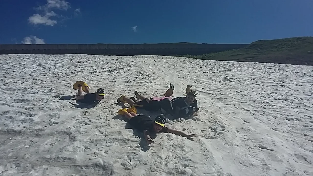 People lying on a snow-covered slope, wearing black T-shirts with googly eyes, yellow shoes, and some with hats, under a blue sky.