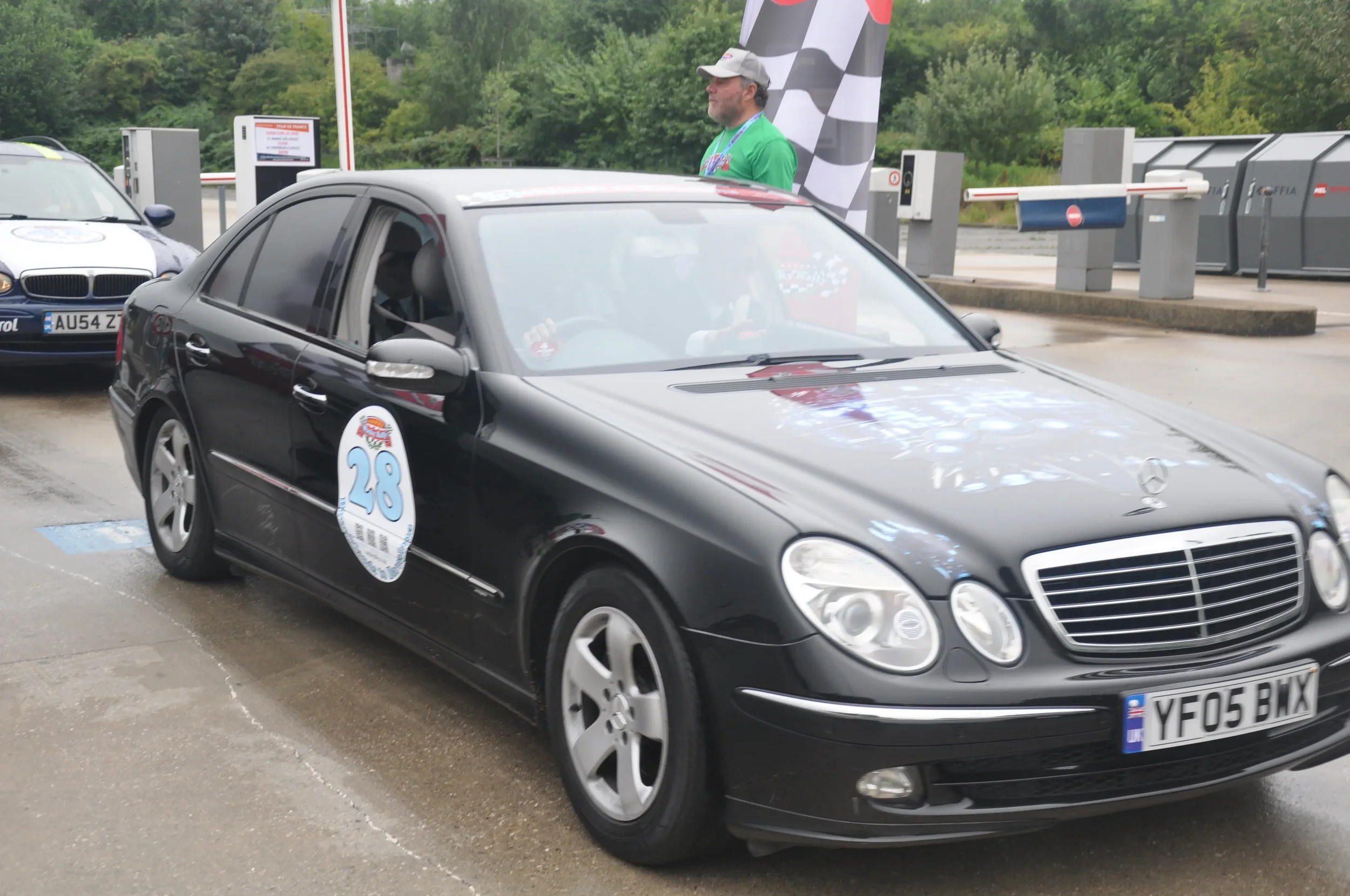 A black Mercedes-Benz with a racing number 28 sticker on the side parked at a race event, with a man standing next to the car and a man in the driver's seat in a racing suit.