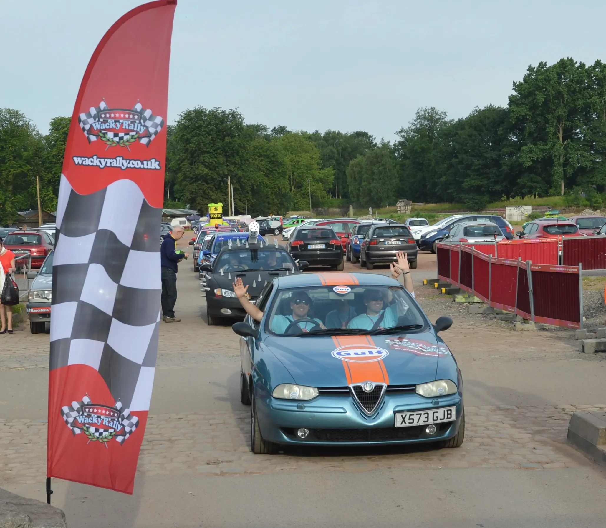 A blue Alfa Romeo car with Gulf racing decals on the hood, driven by two people, is passing a red wacky rally flag in a parking lot filled with cars and onlookers.