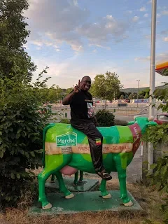 A man sitting on a colorful cow statue outside near a gas station, giving a thumbs-up.