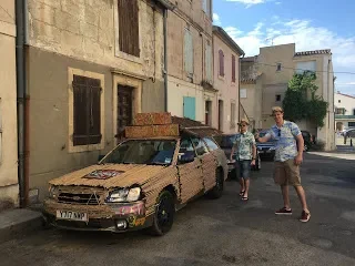 Three people walking past a rusty, abandoned car on a street with old buildings in the background.