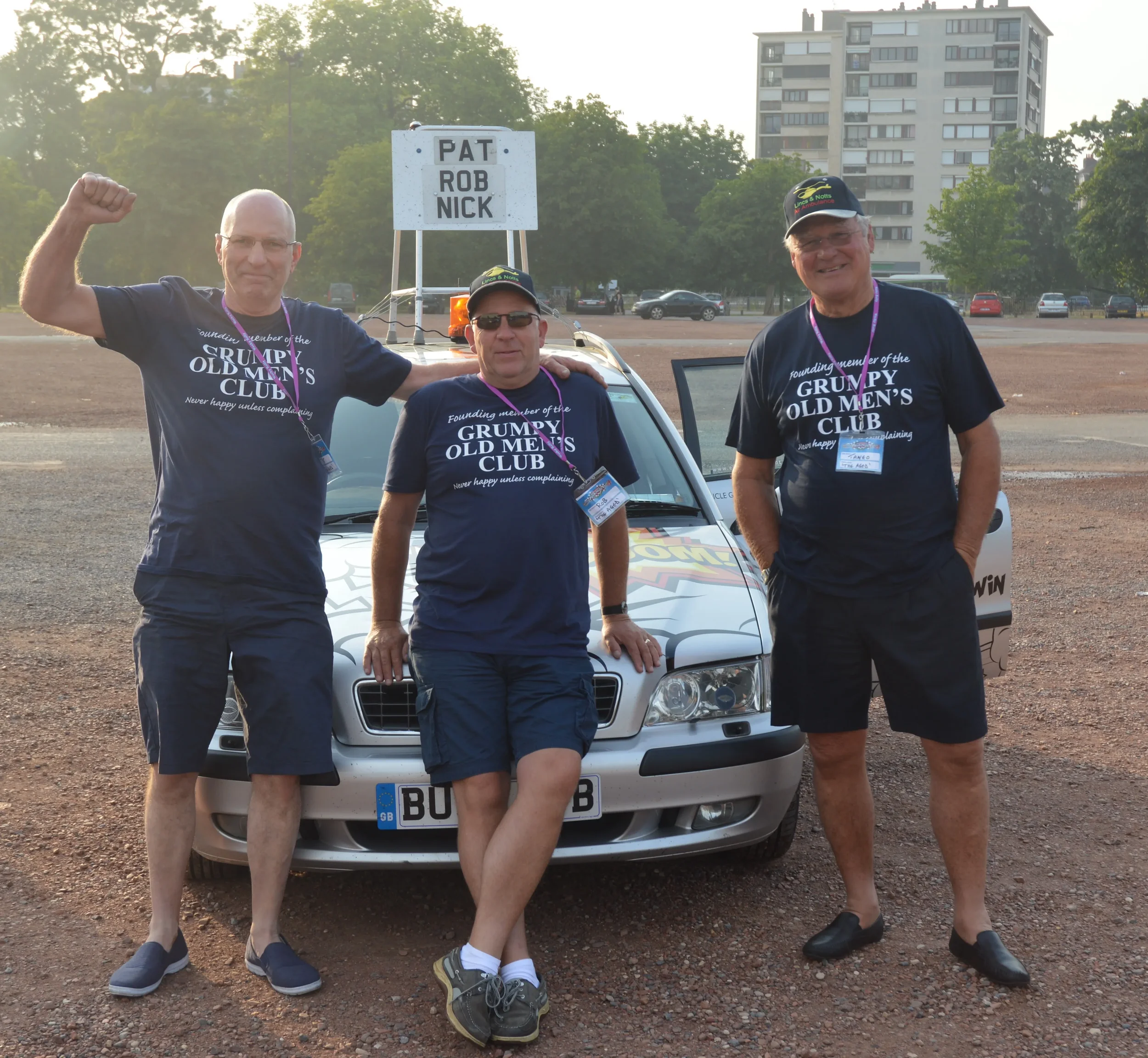 Three middle-aged men standing in front of a decorated silver car in a parking lot, all wearing matching navy T-shirts that say 'Founded member of the GRUMPY OLD MEN'S CLUB', with one man flexing his arm, and all of them wearing hats and name tags.