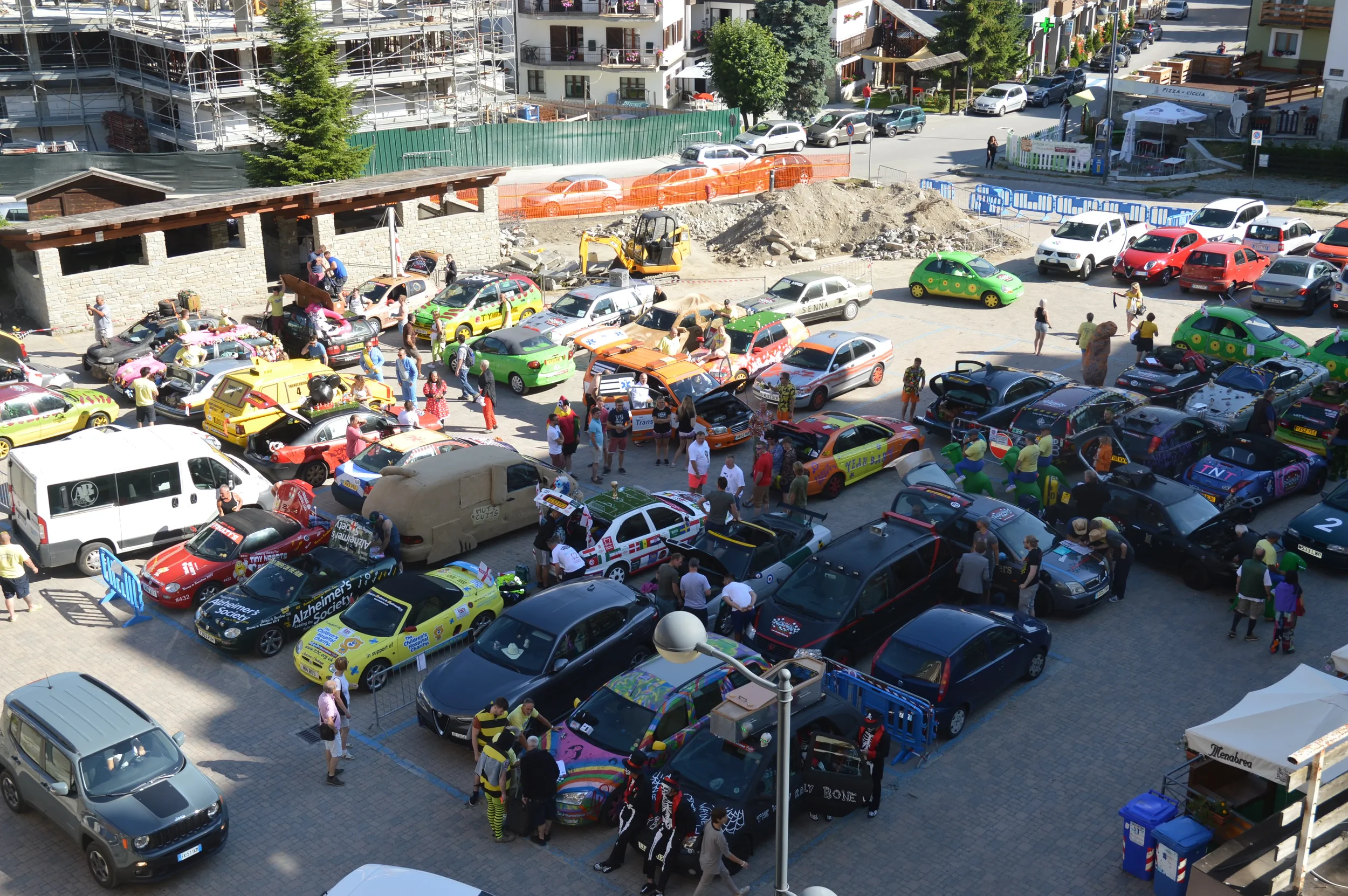 A busy outdoor event with numerous decorated cars, people walking around, and some tents set up in a parking lot surrounded by buildings and construction sites.