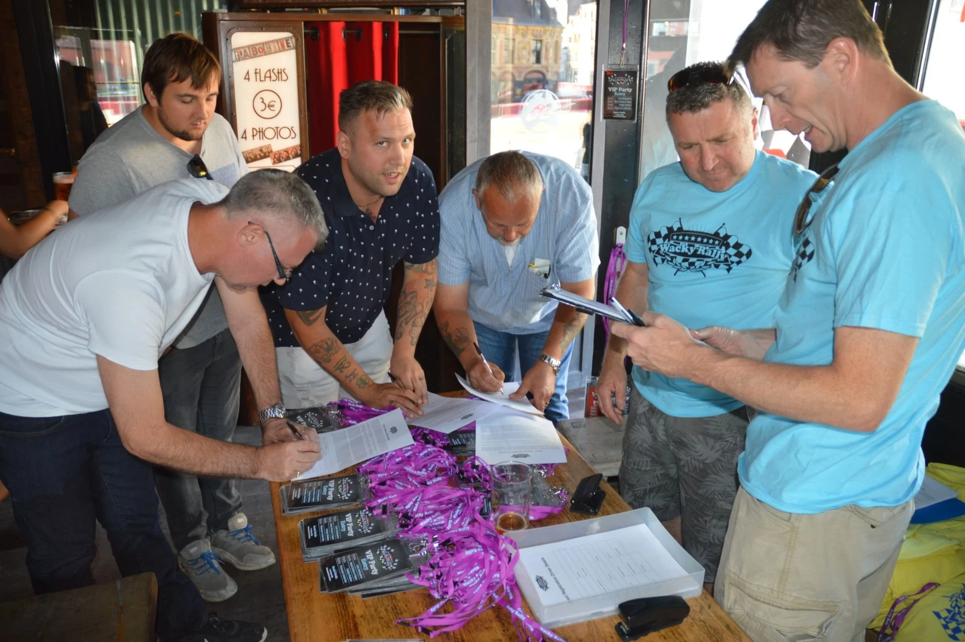 Group of men signing papers at a table with pink lanyards, set inside a bar or restaurant.