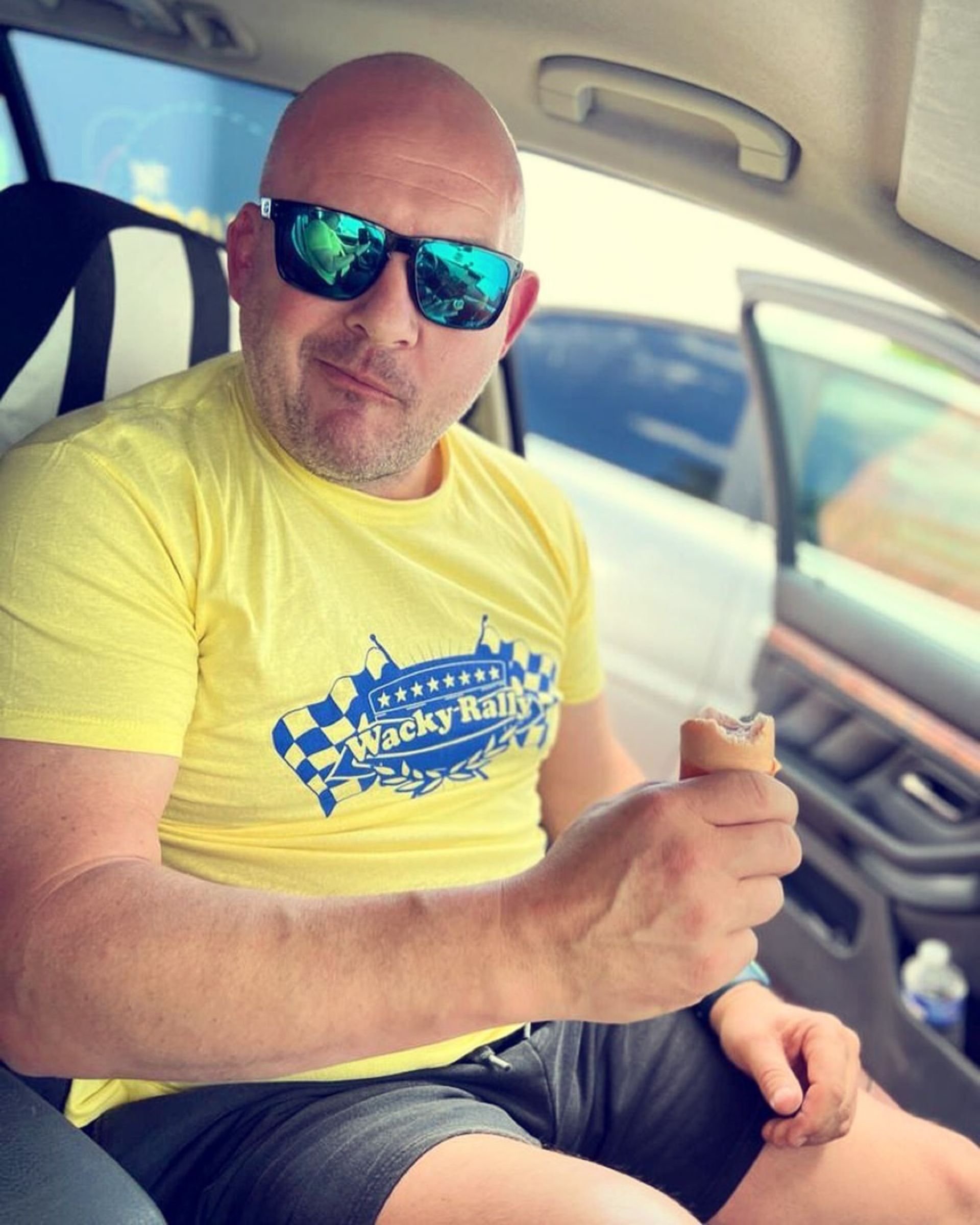 A bald man wearing sunglasses and a yellow T-shirt with a 'Wacky Rally' logo is sitting inside a vehicle, holding an ice cream bar.