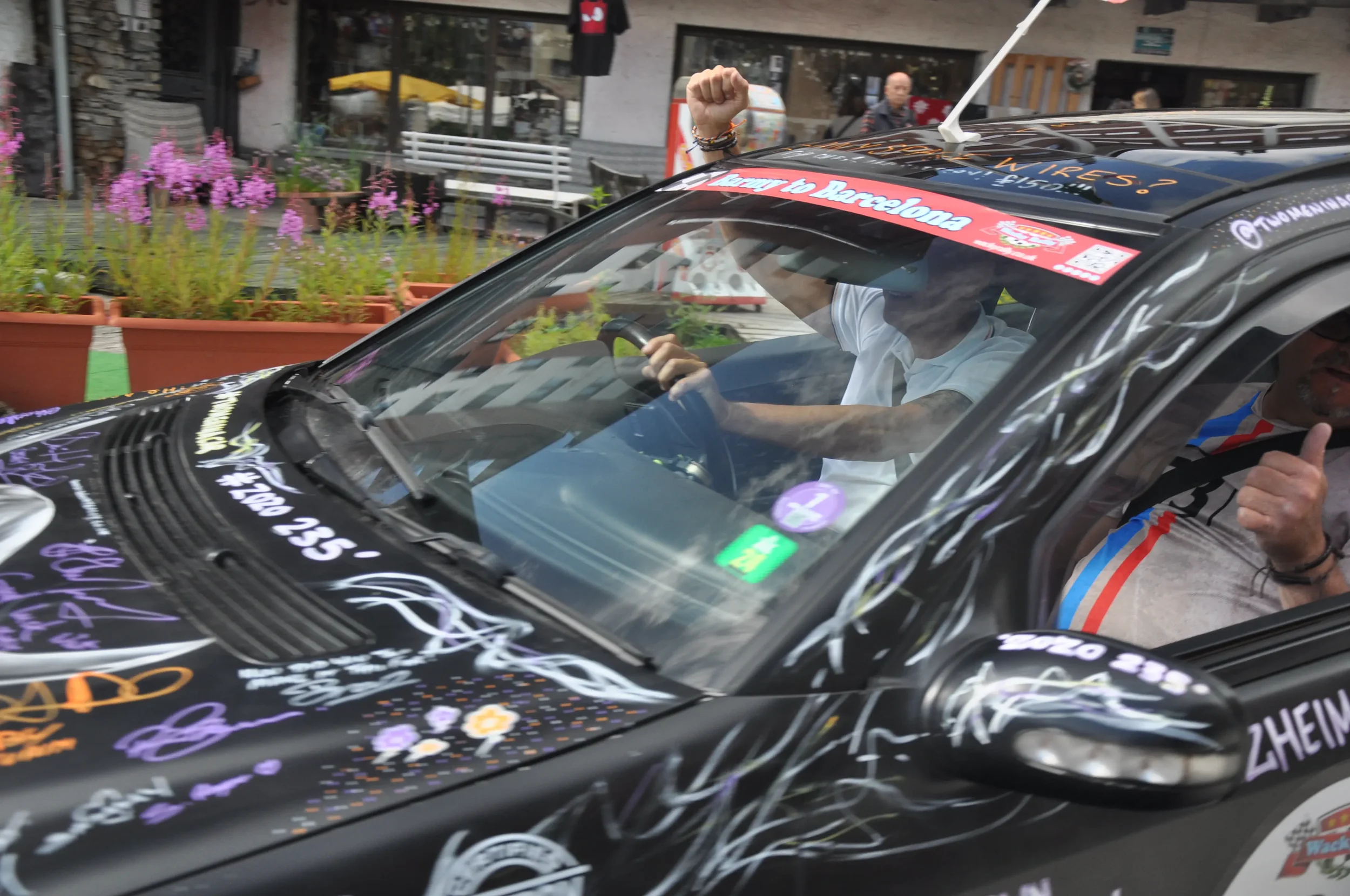 A person in a white shirt driving a decorated black car with purple and white accents during a parade or event. There are pink flowers in pots and a building with people in the background.