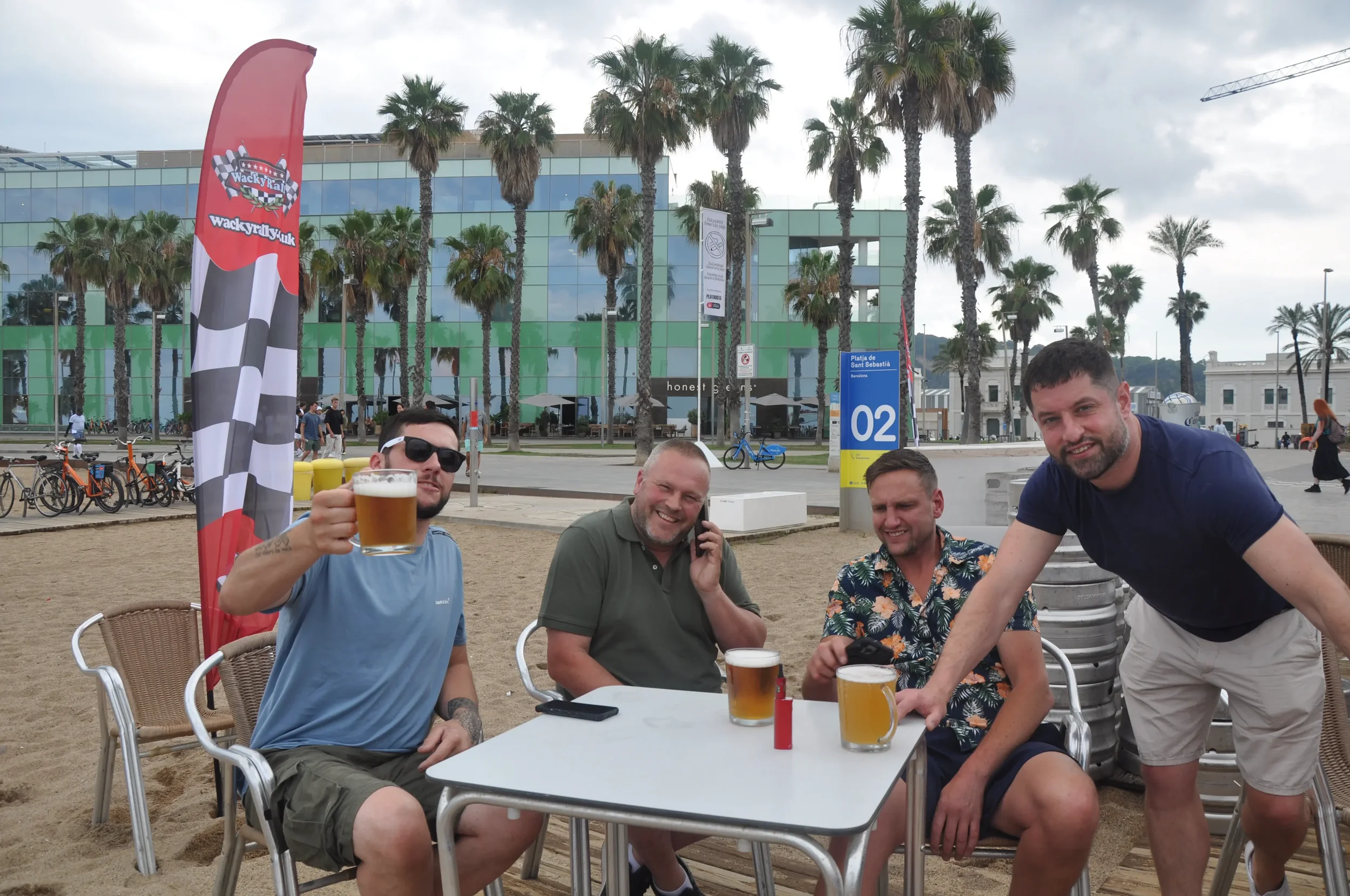 Four men sitting at a table on a sandy beach, enjoying beers, with palm trees and a modern building in the background.