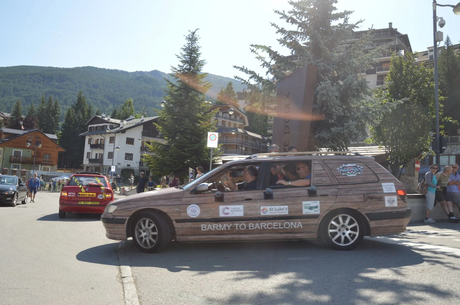 A car wrapped in wood-patterned vinyl with stickers, including one that says 'BARMY TO BARCELONA,' drives through a mountain village street with onlookers on the sidewalk, parked cars, and trees in the background.