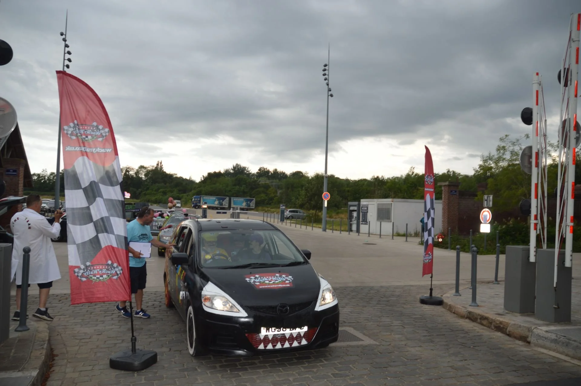 A black car with a race-themed sticker on the hood is at a border crossing or checkpoint, with a person in a blue shirt checking documents and a person in a white coat taking a photo. Flags with checkered patterns mark the area, and there are barrier